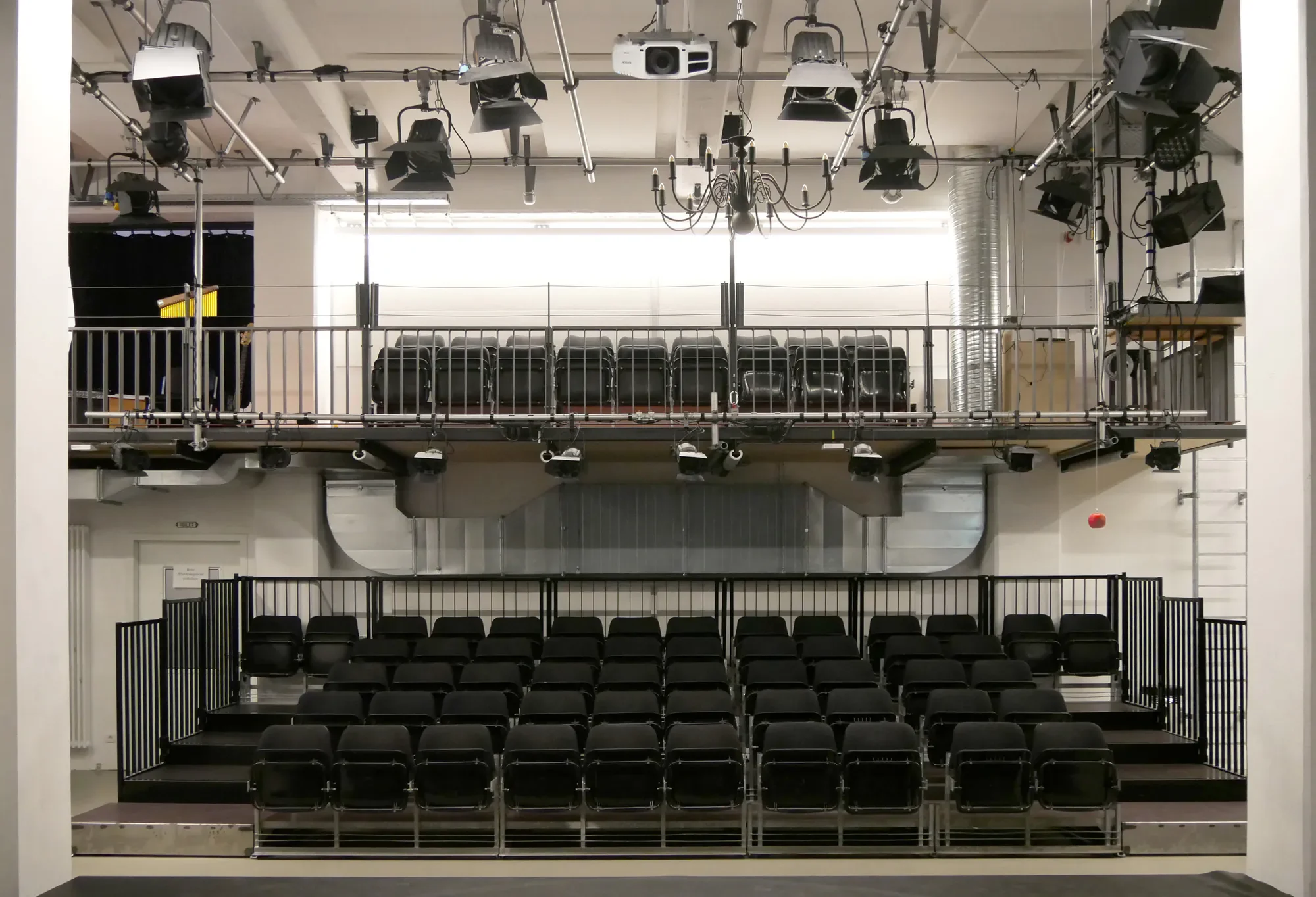 Leeres Theater mit schwarzem Bühnenvorhang, mehreren Reihen schwarzer Sessel, oberer Balkon mit Stuhlreihen, technische Ausstattung einschließlich Scheinwerfer, Projektor und Musikanlage, in einem modernen Veranstaltungsraum.