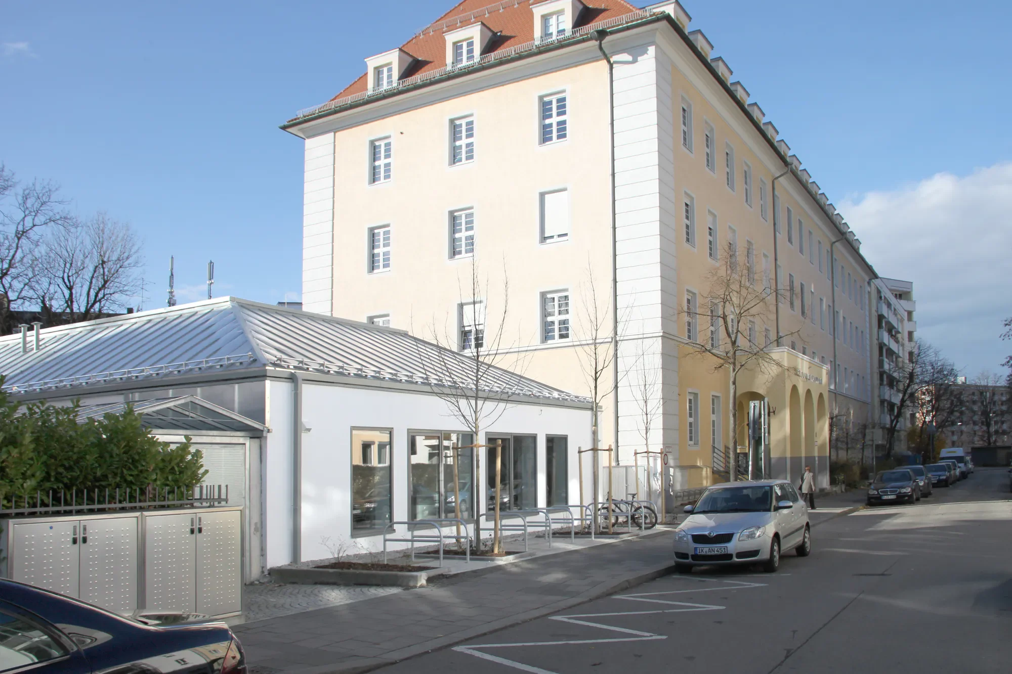 Ein mehrstöckiges Gebäude mit weißen und gelben Wänden und roten Dächern. Im Vordergrund befinden sich geparkte Autos, eine Bushaltestelle und Bäume ohne Laub.