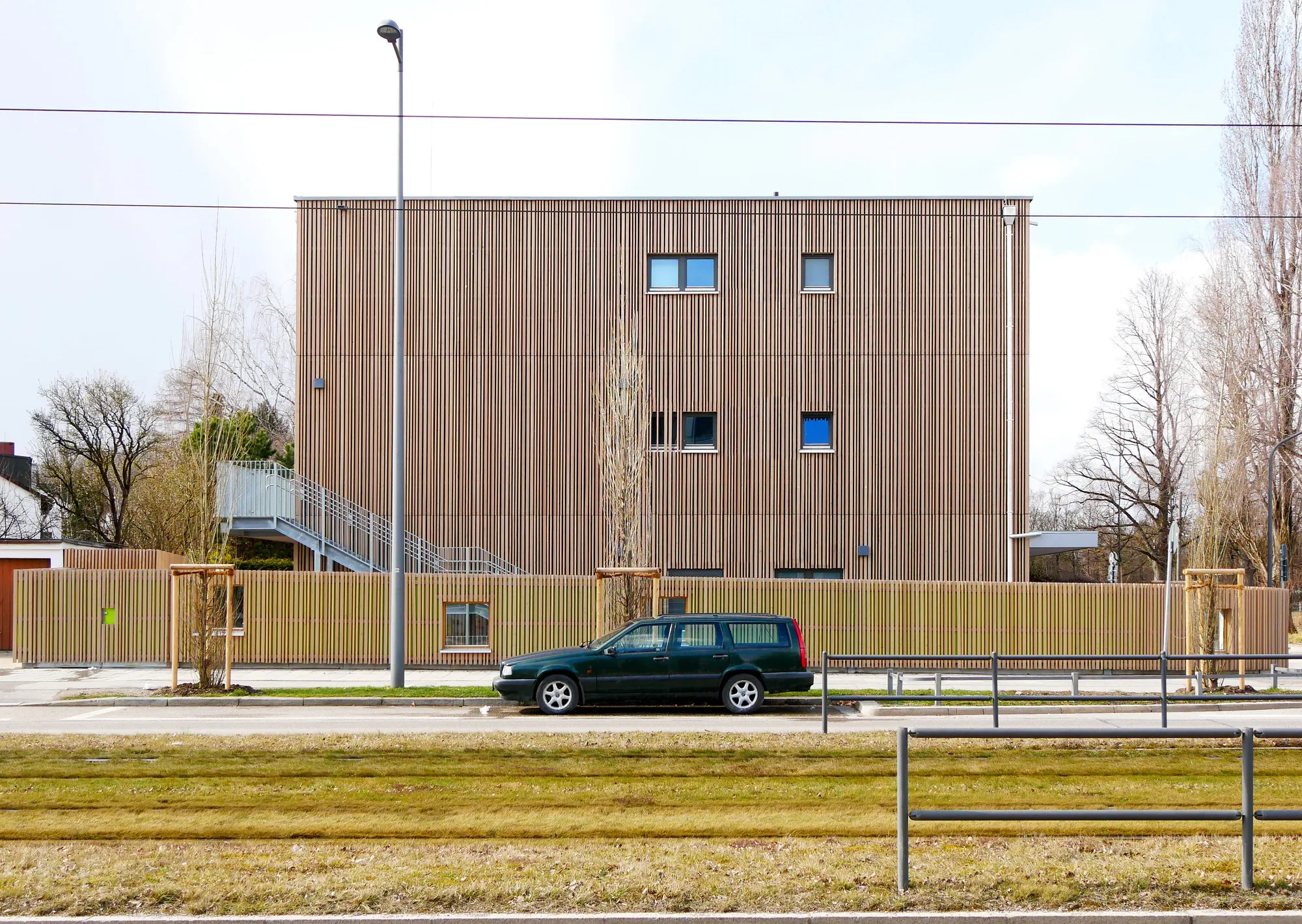 Ein modernes, mehrstöckiges Gebäude mit einer Holzfassade, mehreren kleinen Fenstern, einem Zaun im Vordergrund, einem schwarzen Fahrzeug auf der Straße und Bäumen im Hintergrund.