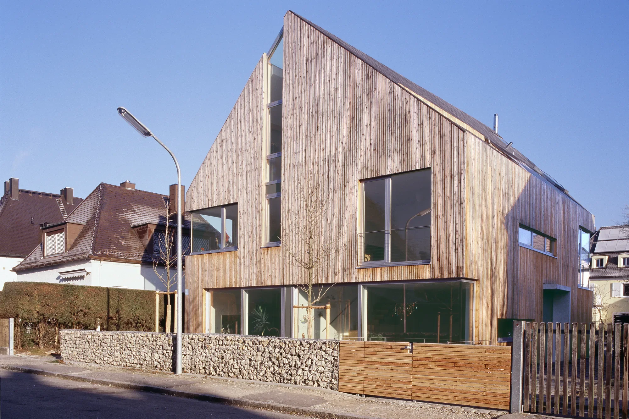 Modernes dreistöckiges Haus mit Holzfassade, große Fenster, Zaun und Straßenlaterne im Vordergrund, benachbarte Häuser und blauer Himmel im Hintergrund.