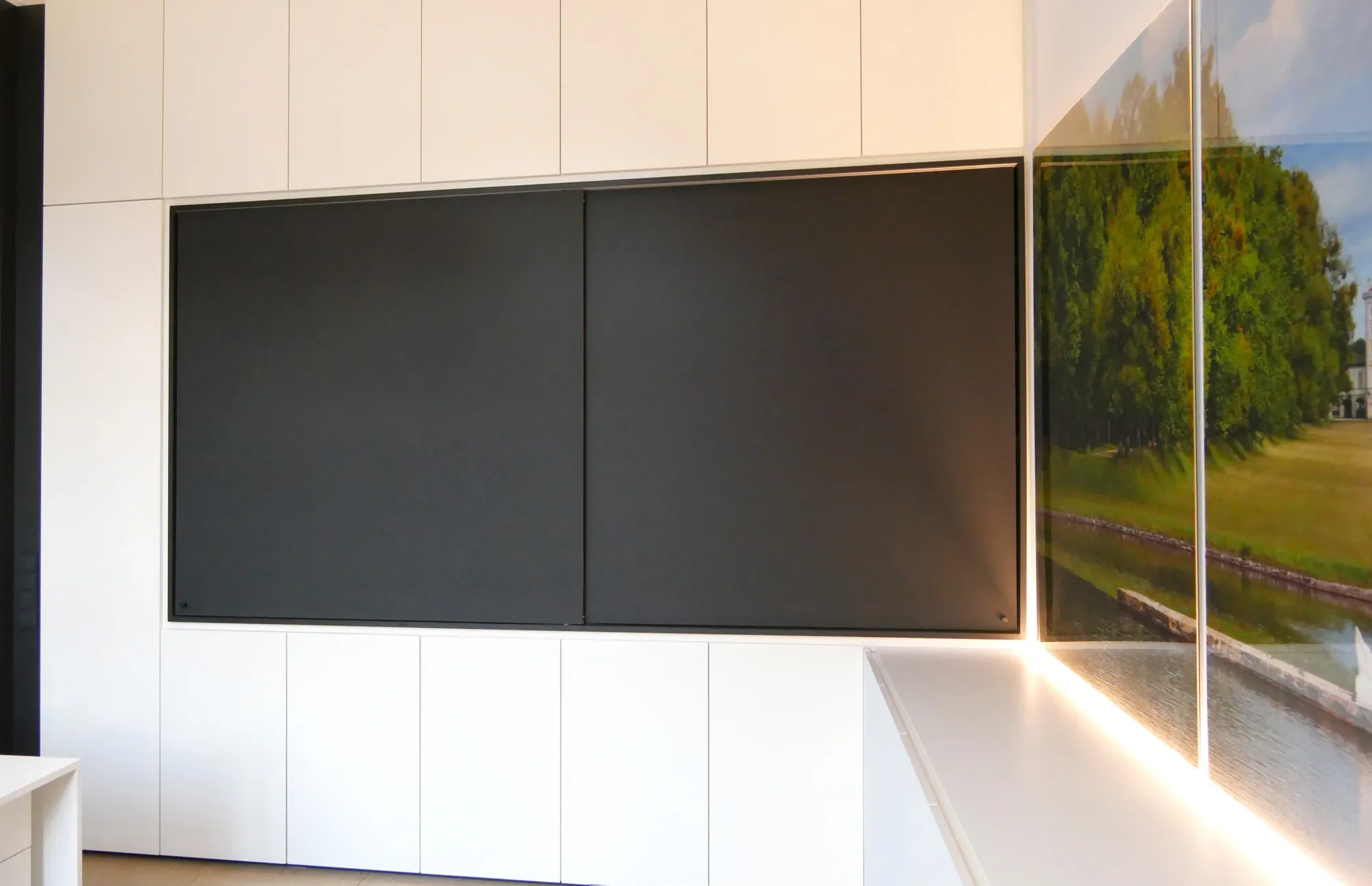Moderne, weiße Wand mit eingebauten Schränken und einem großen Fenster, das eine grüne Parklandschaft mit Bäumen und einem Wasserloch zeigt.