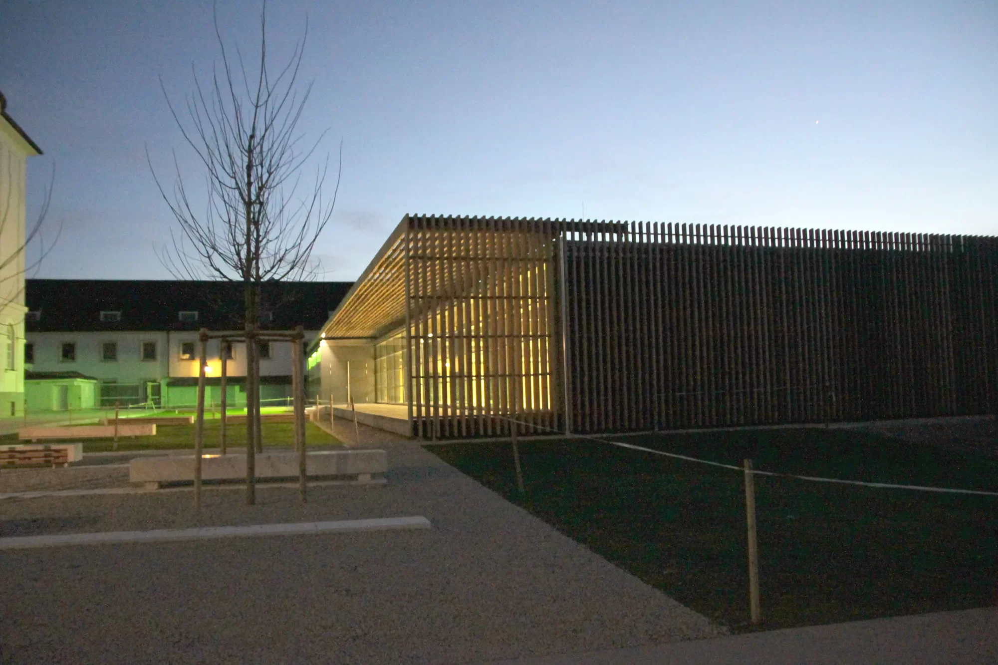 Modernes Gebäude mit einer Holzfassade und großen Fenstern in einem Park bei Nacht, im Hintergrund sind Bäume und andere Gebäude sichtbar.