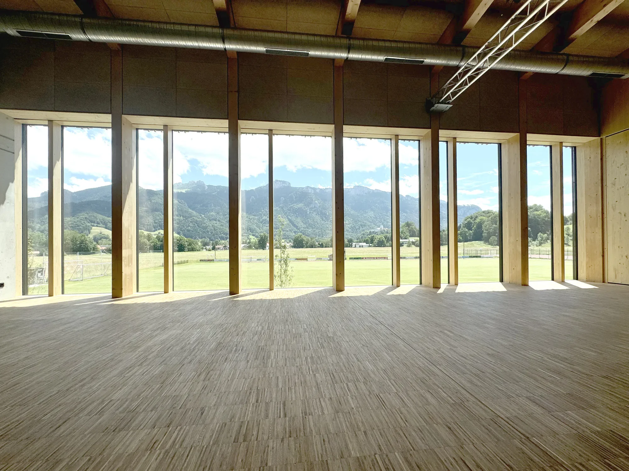 Innenaufnahme eines Raumes mit großen Fensterfronten und Blick auf Berge im Hintergrund, unfertiger Bau mit sichtbaren Holzrahmen und Deckenrohren.