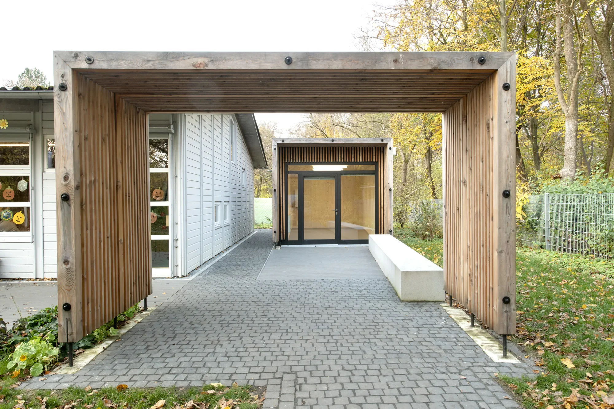 Moderner Eingang zu einer Einrichtung mit Holzrahmen und Glasfront, umgeben von Bäumen im Herbst, gepflasterter Weg führt zur Tür.