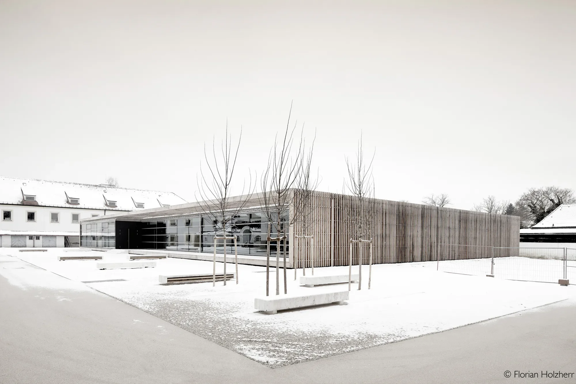 Ein moderner Gebäudekomplex mit einer Holzfassade und großen Glasfenstern, umgeben von schneebedecktem Boden und Bäumen ohne Blätter.