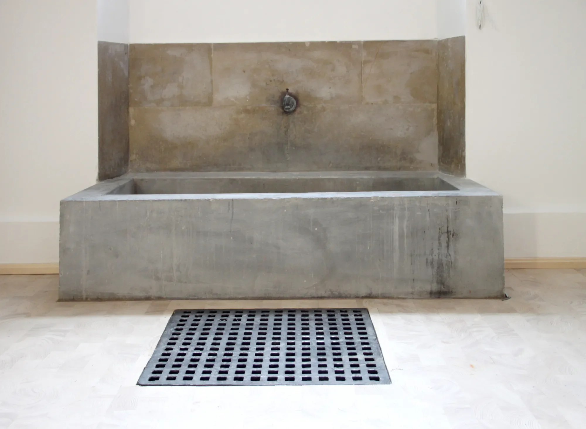 Ein Waschraum mit grauem Steinwaschbecken, einem Abfluss im Boden und einem Metallgitter als Abfluss. Die Wände sind hell, der Boden ist beige.