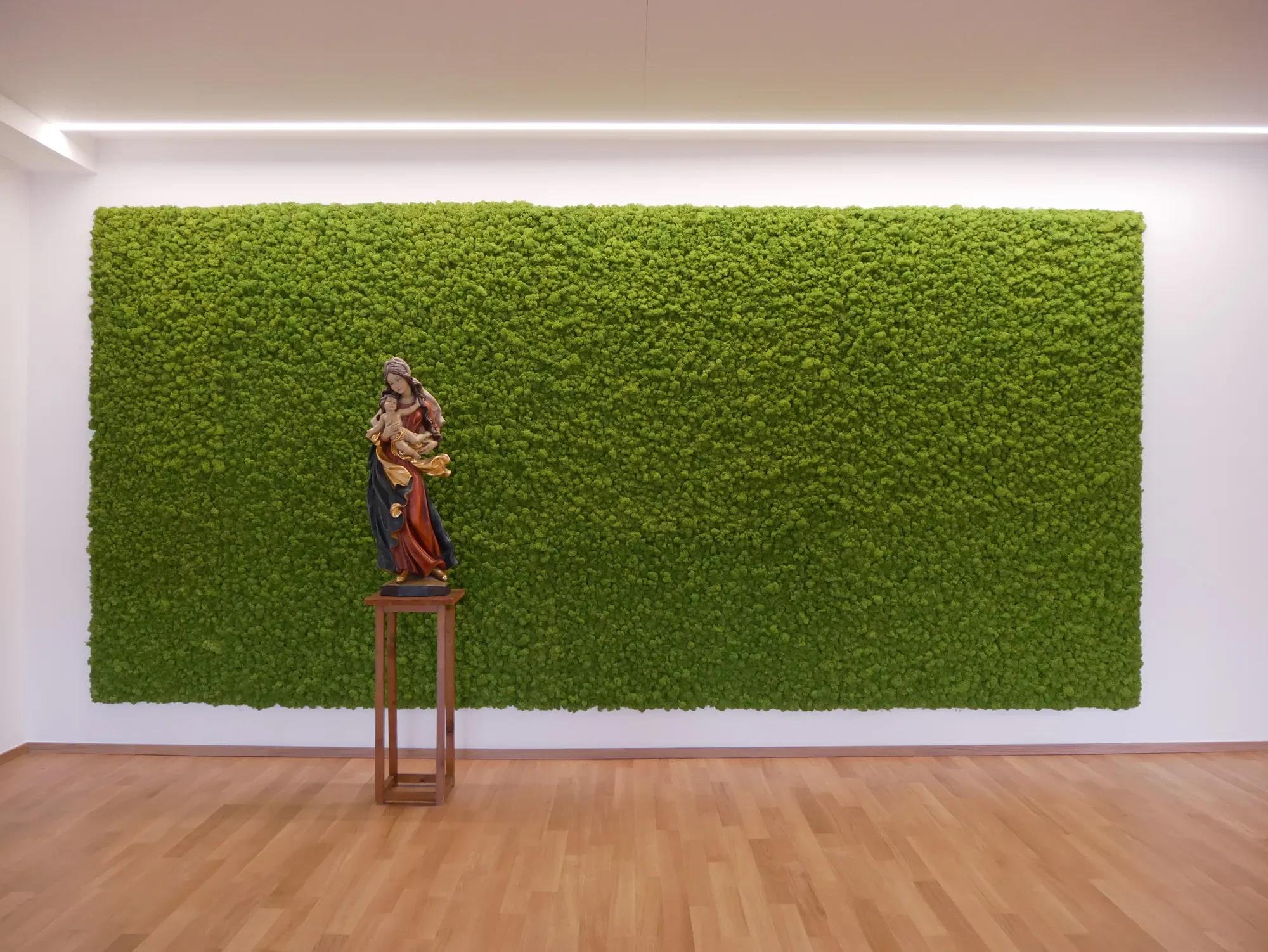 Kunstvolle Sakralfigur vor grünem Moos Wand in einem weißen Raum, auf einem kleinen Holztisch