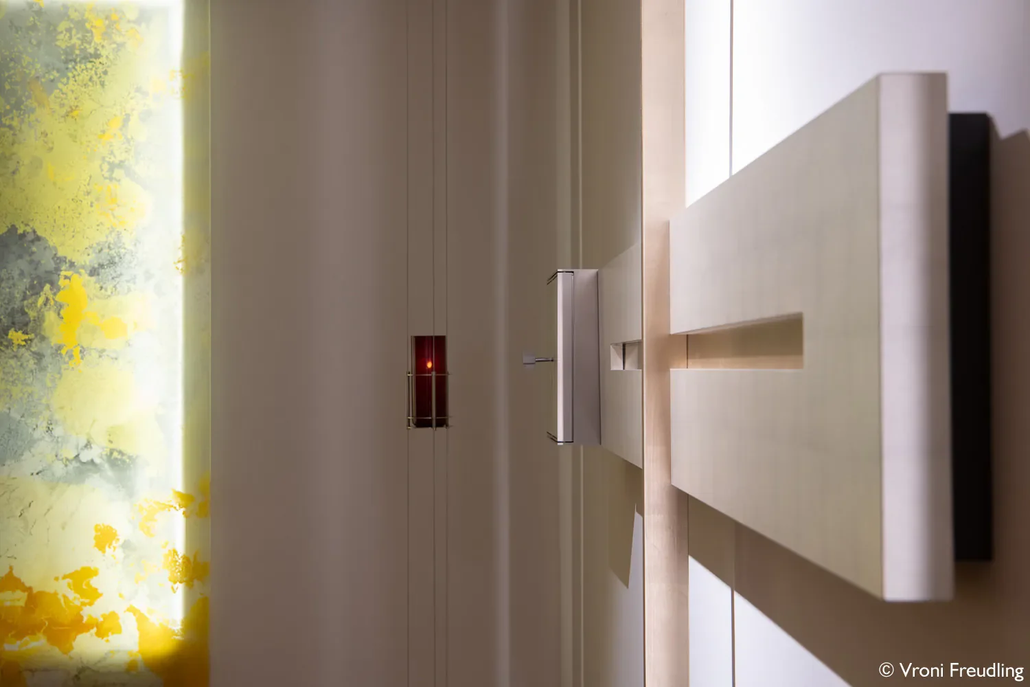 Innenaufnahme eines modernen, minimalistischen Raums mit hellen Wänden, einem hellen Boden und einem großen, abstrakten Wandbild mit gelben und grünen Akzenten auf der linken Seite. Im Vordergrund befindet sich eine lange, schmale Holzbank oder -tisch. In der Mitte des Bildes ist eine kleine, rote Lichtquelle oder ein elektronisches Gerät sichtbar.