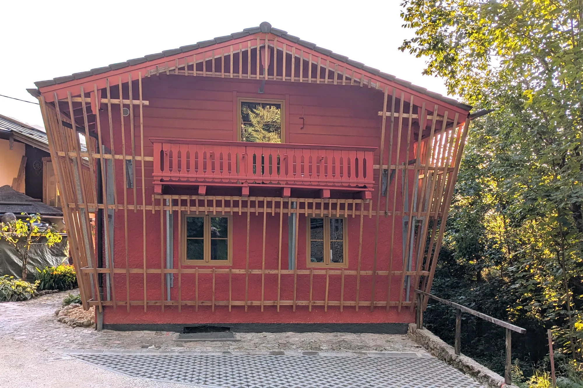 Ein Haus in Bau mit roter Fassade und Holzrahmen, das im oberen Teil noch unvollständig ist, mit einer Balkon- und Fenstermontage. Es ist von Bäumen umgeben und es scheint, dass gerade Bauarbeiten stattfinden.