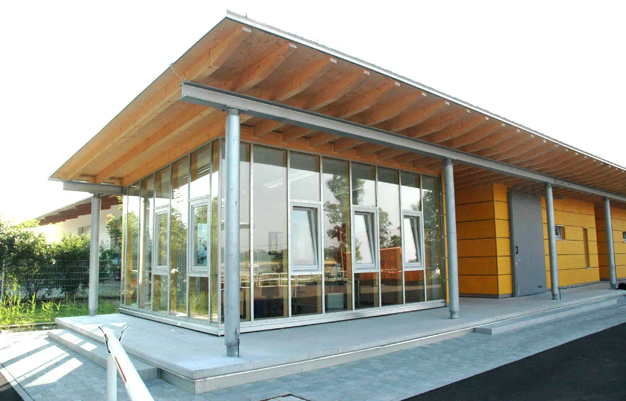 Modernes Gebäude mit Holz und Glasfassade, große Fenster, geneigtes Dach, Metallstützen, umgeben von Garten und Gehweg.