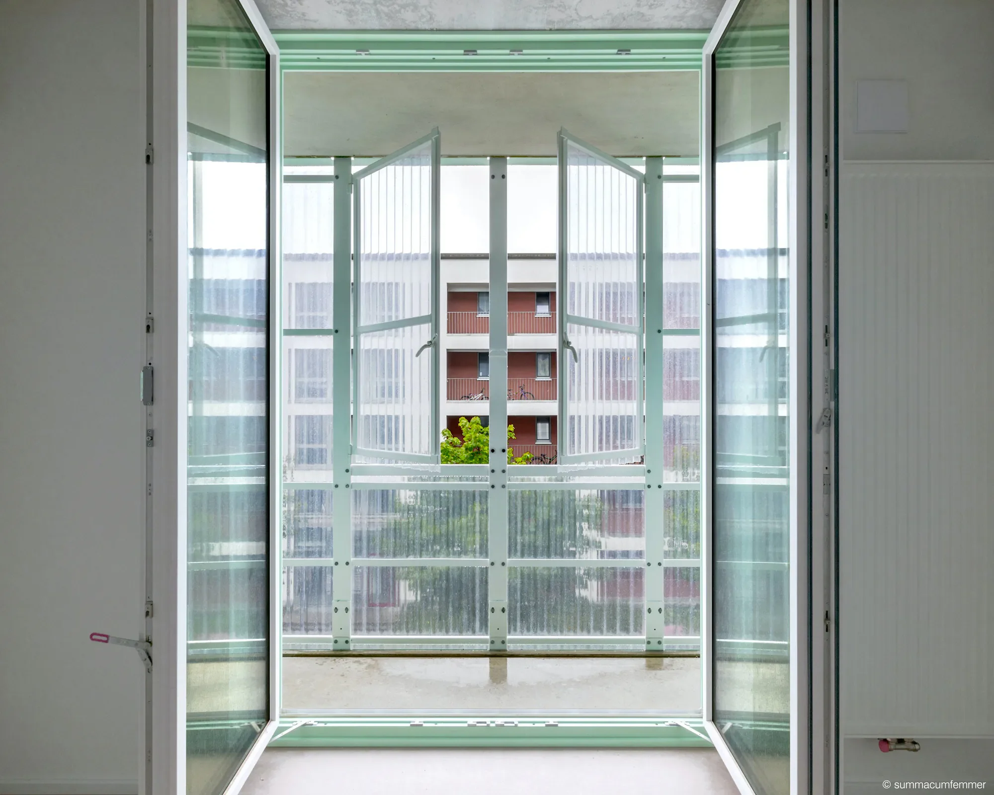 Offene Fenster eines Gebäudes mit Blick auf eine Mehrfamilienwohnanlage. Das Bild zeigt die Innenansicht mit Blick nach draußen auf die Balkone und Fassaden der Nachbargebäude.