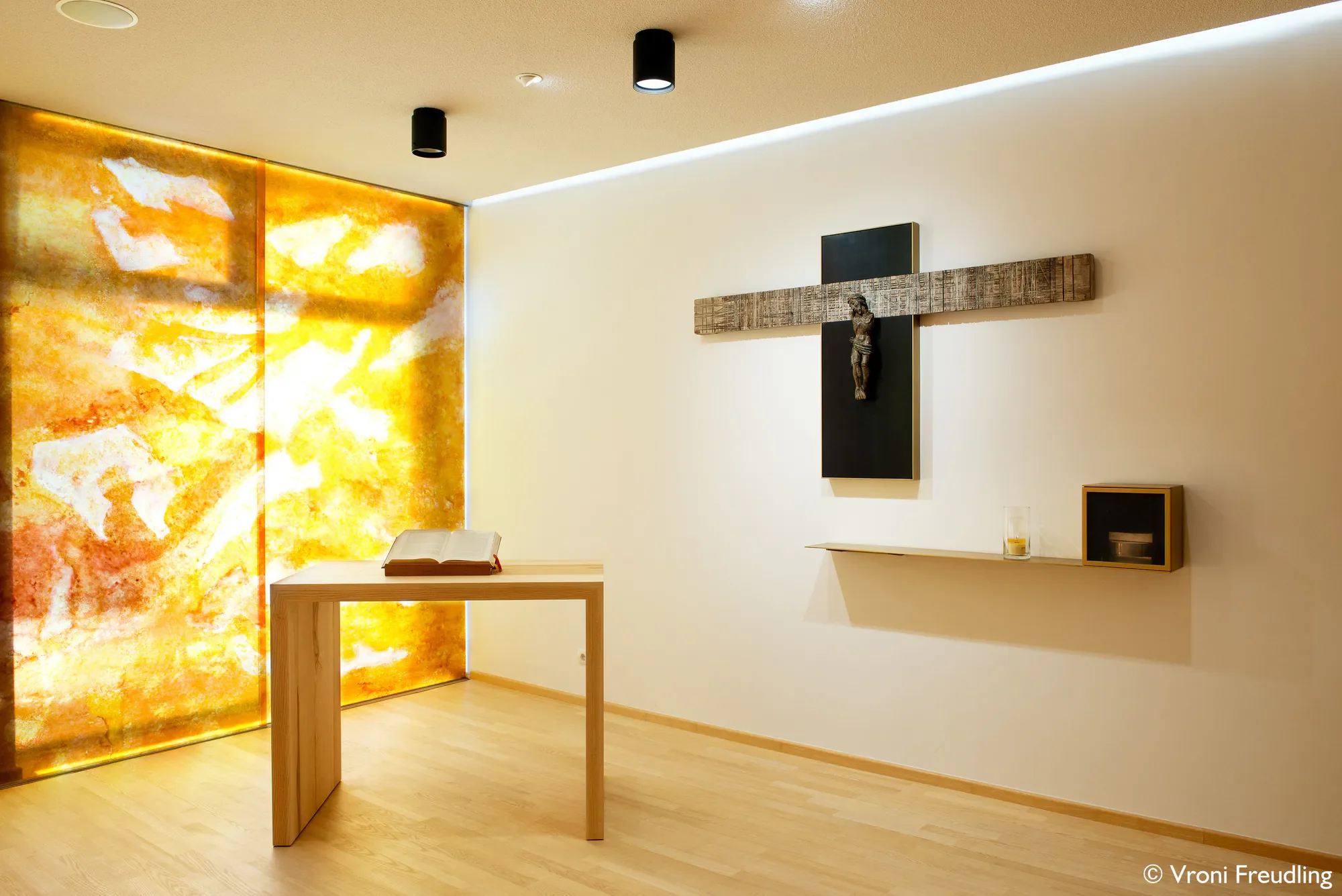 Ein christliches Altarbild mit einem Kreuz, einem Holzstamm und einer Skulptur an der Wand, neben einem Bücherregal und einer Tischdekoration, in einem hellen Raum mit Holzfußboden.