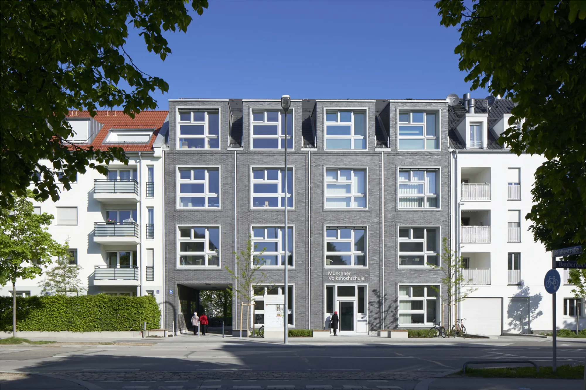 Mehrstöckiges modernes Wohngebäude mit weißen und grauen Fassaden, Bäumen im Vordergrund, blauer Himmel, Straße und Fußgänger davor, Beschilderung für Fahrradweg