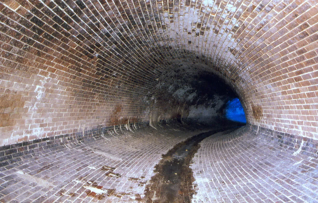 Blick in einen breiten, bogenförmigen BS-Tunnel mit Ziegelsteinmauerung, Wasserpfütze auf dem Boden und einem schwachen blauen Licht am Ende.