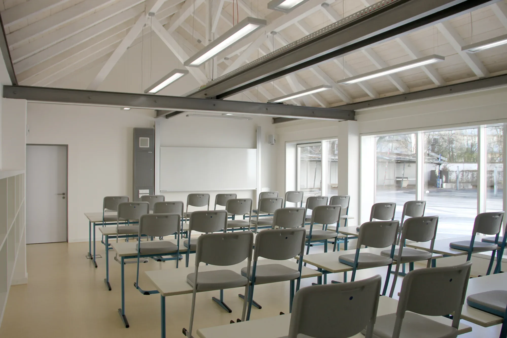 Leeres Klassenzimmer mit weißen Stühlen auf Tischen, große Fenster, strukturierte Decke mit Beleuchtung, Whiteboard und Sitzgelegenheiten.