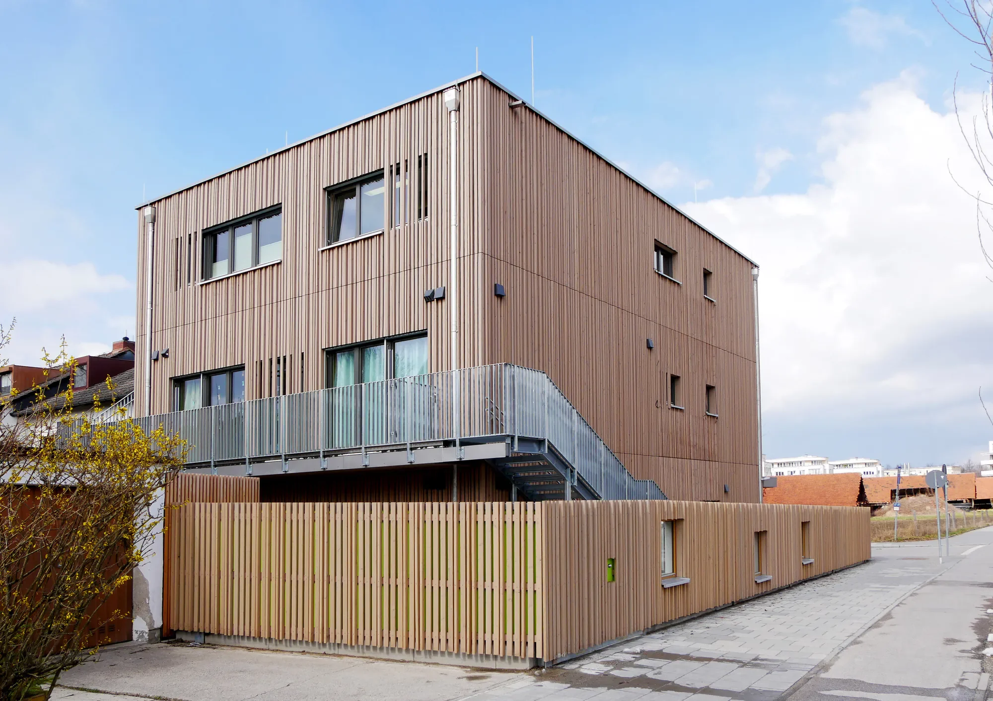 Mehrstöckiges modernes Gebäude mit Holzfassade und Balkon, umgeben von Bürgersteig und niedrigen Zaun, bei bewölktem Himmel.