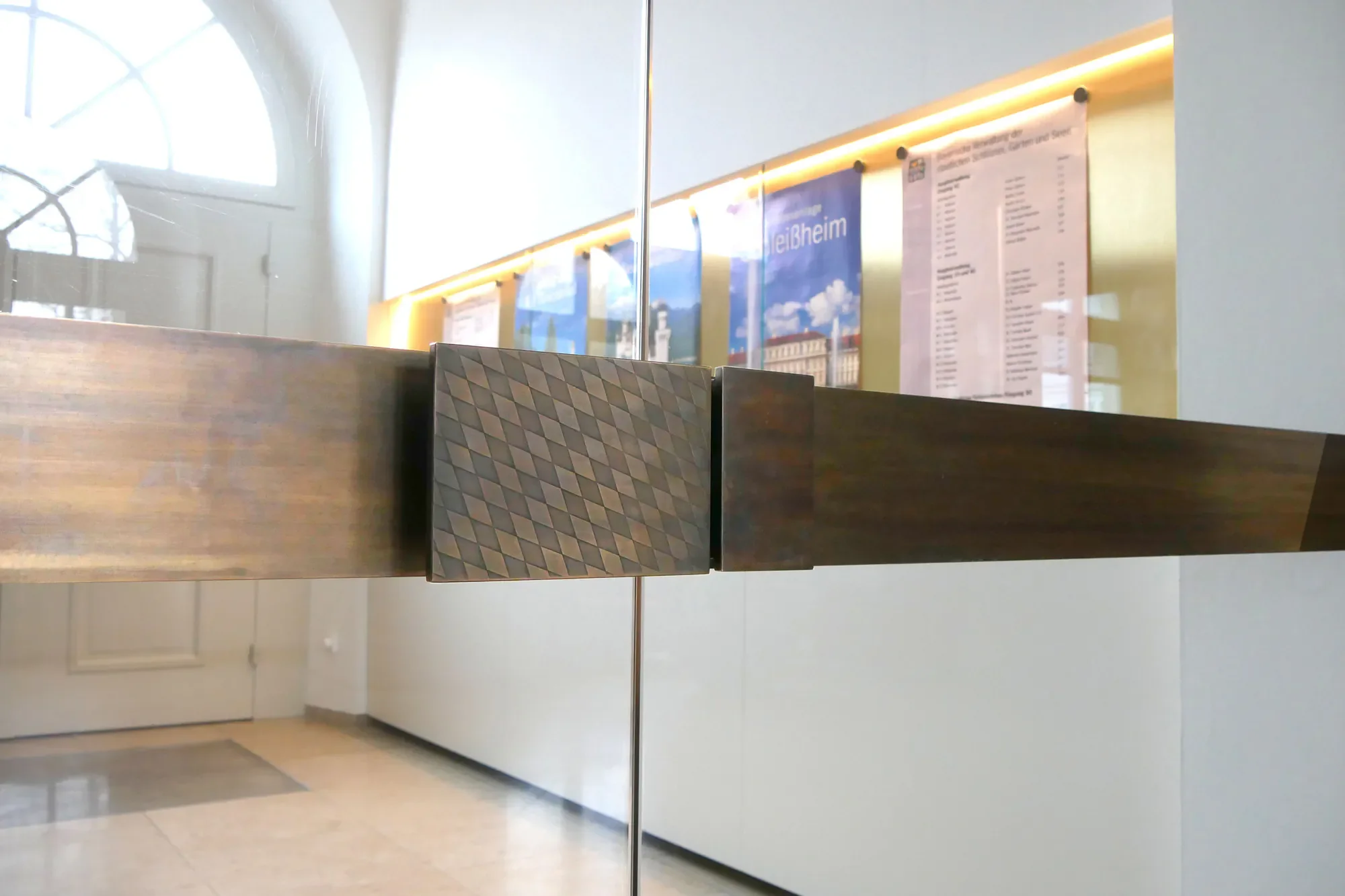 Innenaufnahme eines Warteschlangenbereichs mit einem durchsichtigen Glasfenster, einer goldfarbenen Wand mit einem Aushang und einem Holztresen im Vordergrund.