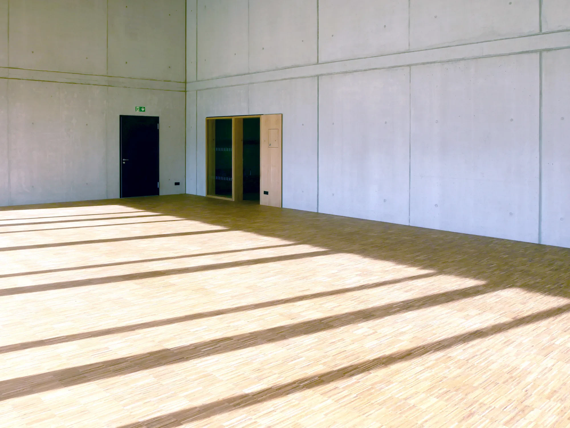 Ein Raum mit gelblichem Holzboden, weichen Schattenmuster und weißen Betonwänden. Es gibt eine schwarze Tür und eine gelbe Glasvitrine an der Wand.