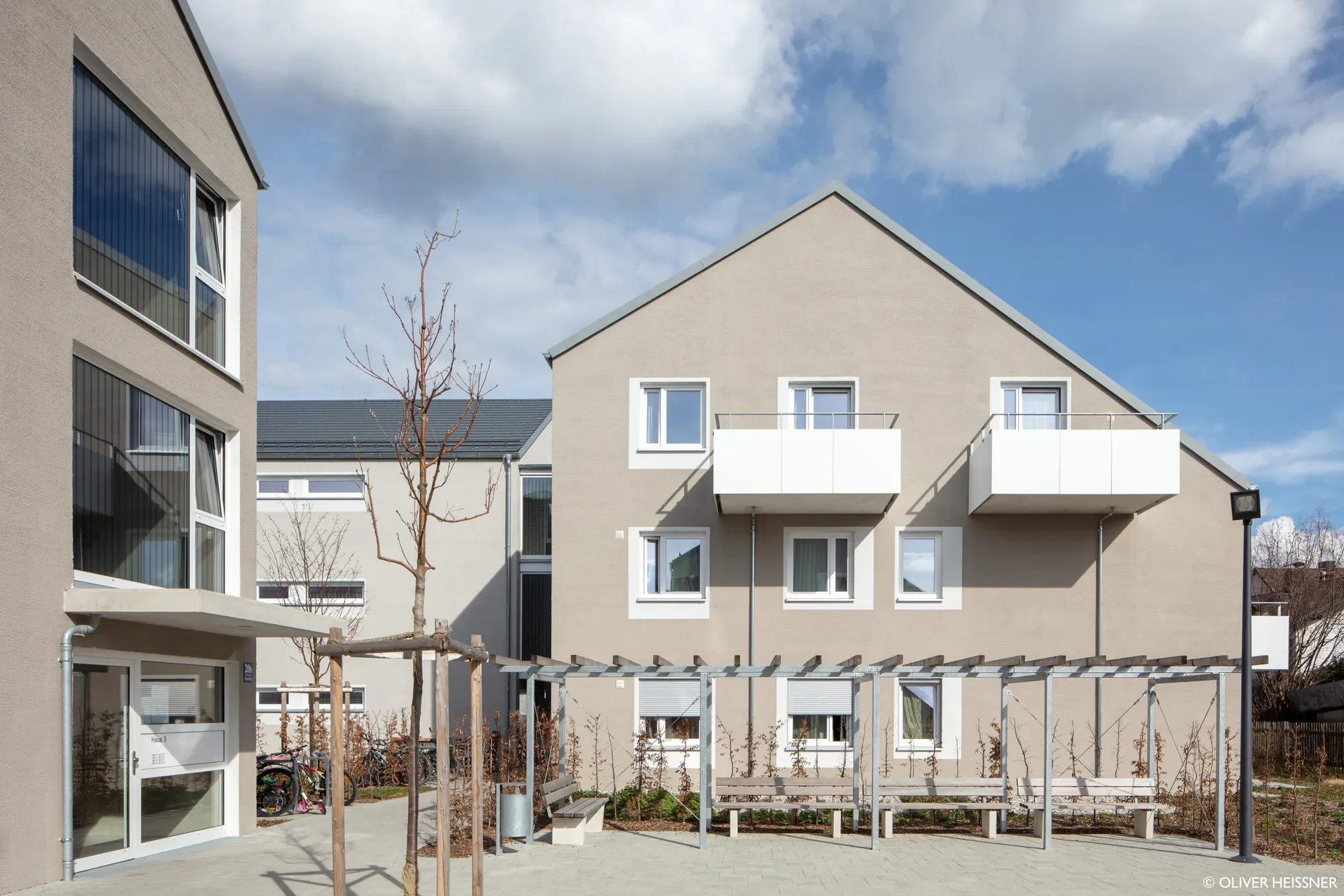 Mehrfamilienhaus mit braunem Pultdach und weißen Balkonen, Vordergrund mit Sitzbänken, Bäumen und Fahrradständer, blauer Himmel mit Wolken.