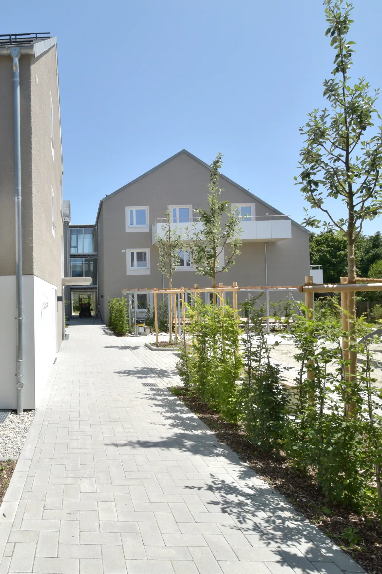 Ein Spazierweg mit Pflastersteinen, grüne Büsche auf der rechten Seite und ein modernes Mehrfamilienhaus im Hintergrund mit mehreren Balkonen und weißen Fenstern. Es ist ein sonniger Tag mit klarem blauen Himmel.