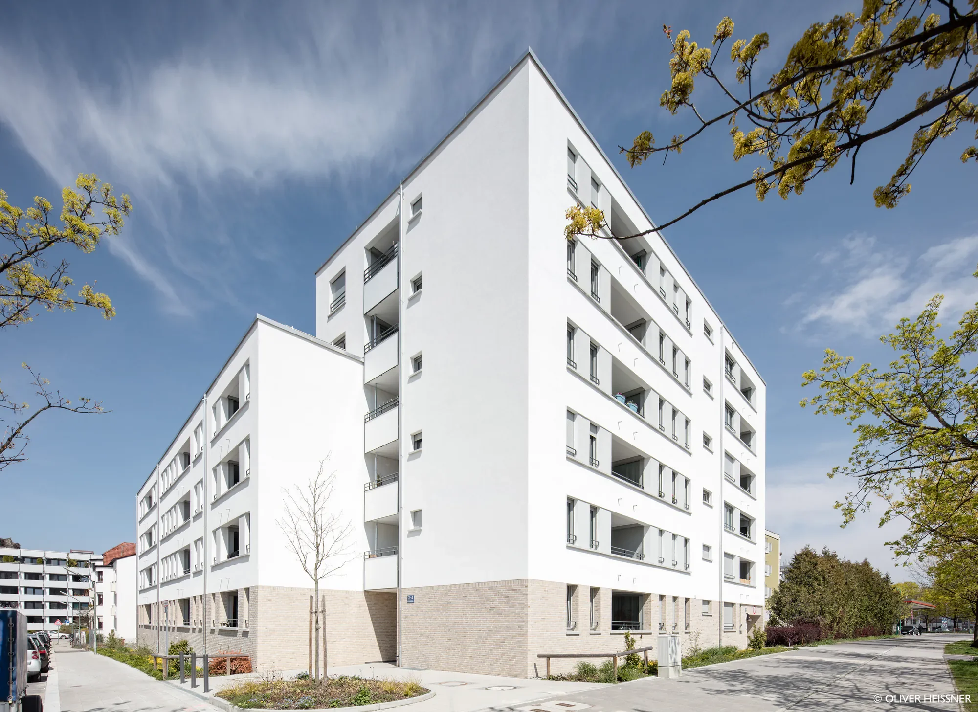 Modernes weißes Mehrfamilienhaus mit mehreren Balkonen, umgeben von Bäumen, bei sonnigem Himmel.