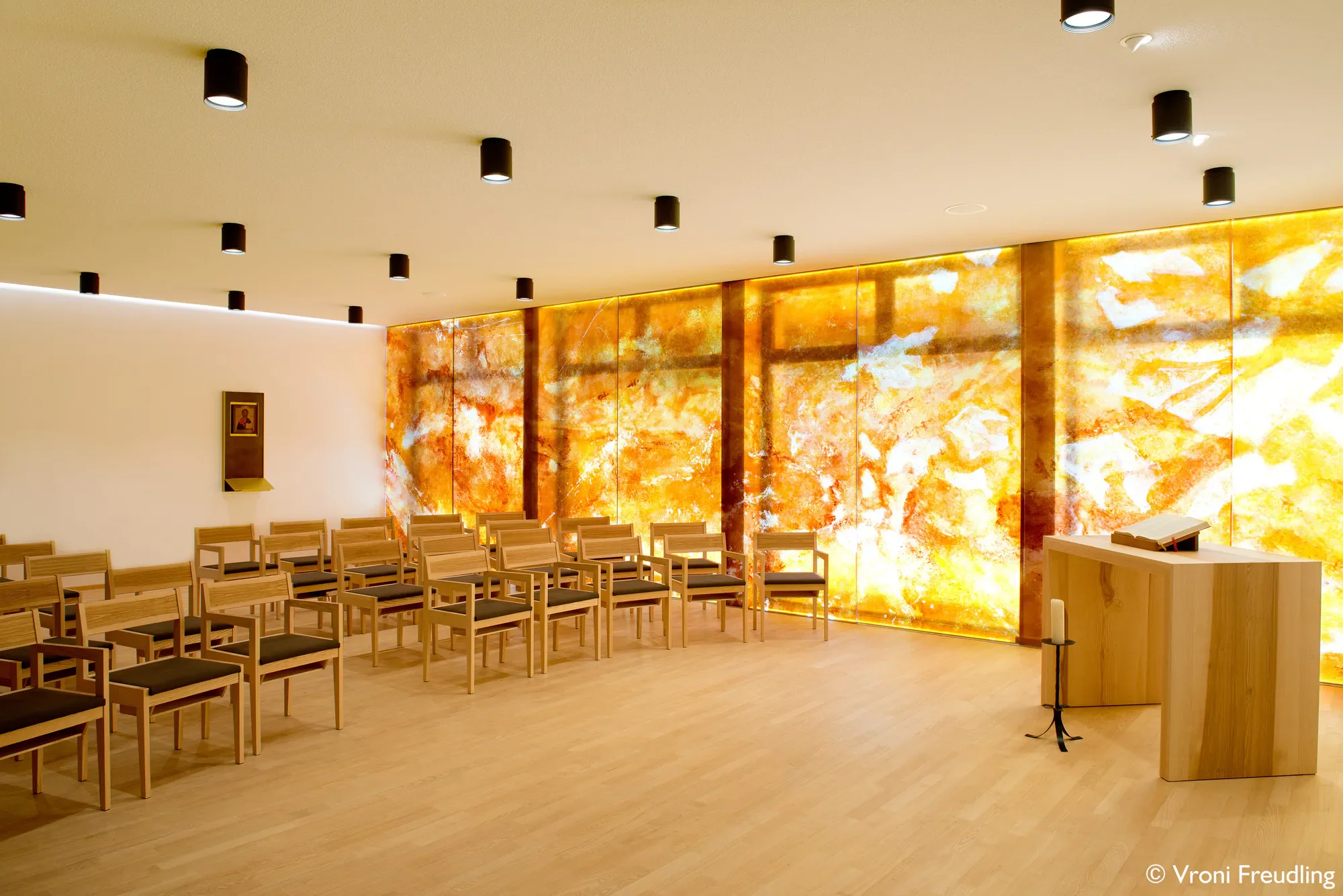 Ein Saal mit mehreren Reihen aus Holzstühlen, einem Altar und einer leuchtenden Wand aus orangefarbenem Marmor.