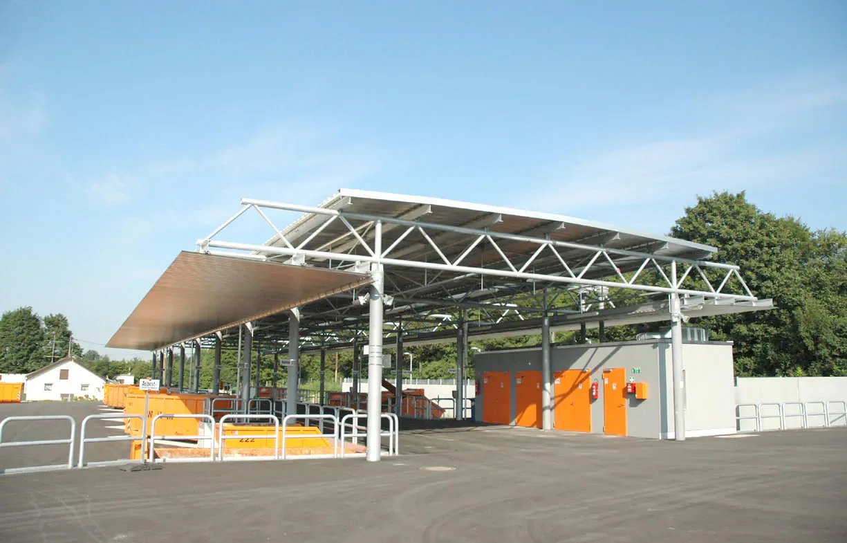 Ein moderner, offener Müllentsorgungsbereich mit Sonnenkollektoren auf dem Dach, orangefarbigen Containern und Sicherheitszäunen, im Hintergrund Bäume und ein blauer Himmel.