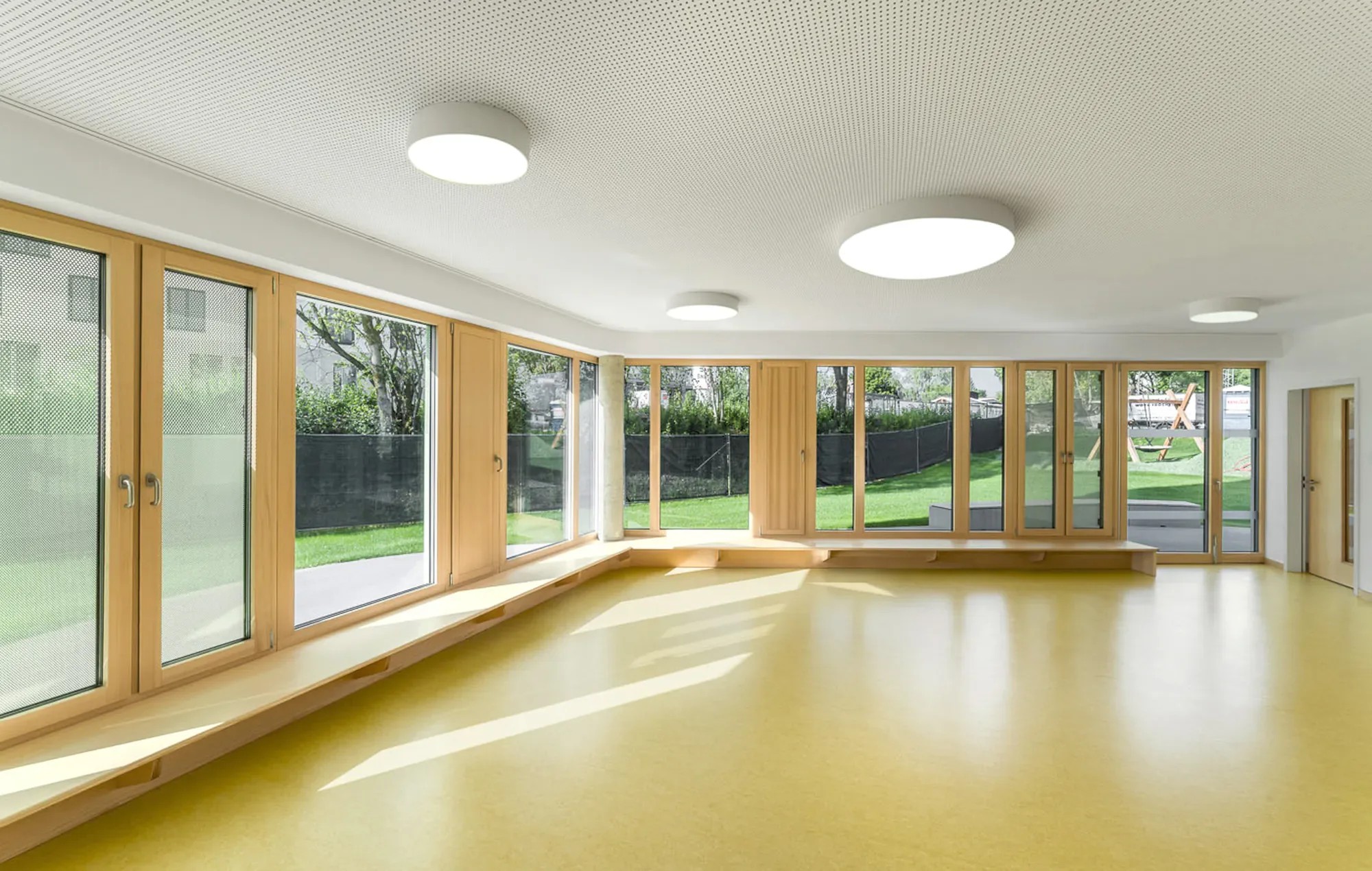Ein heller Raum mit großen Fensterflächen und gelbem Boden, Blick nach draußen auf eine grüne Wiese mit Bäumen, innerraum hat Deckenleuchten und eine Holzbank entlang der Fenster.