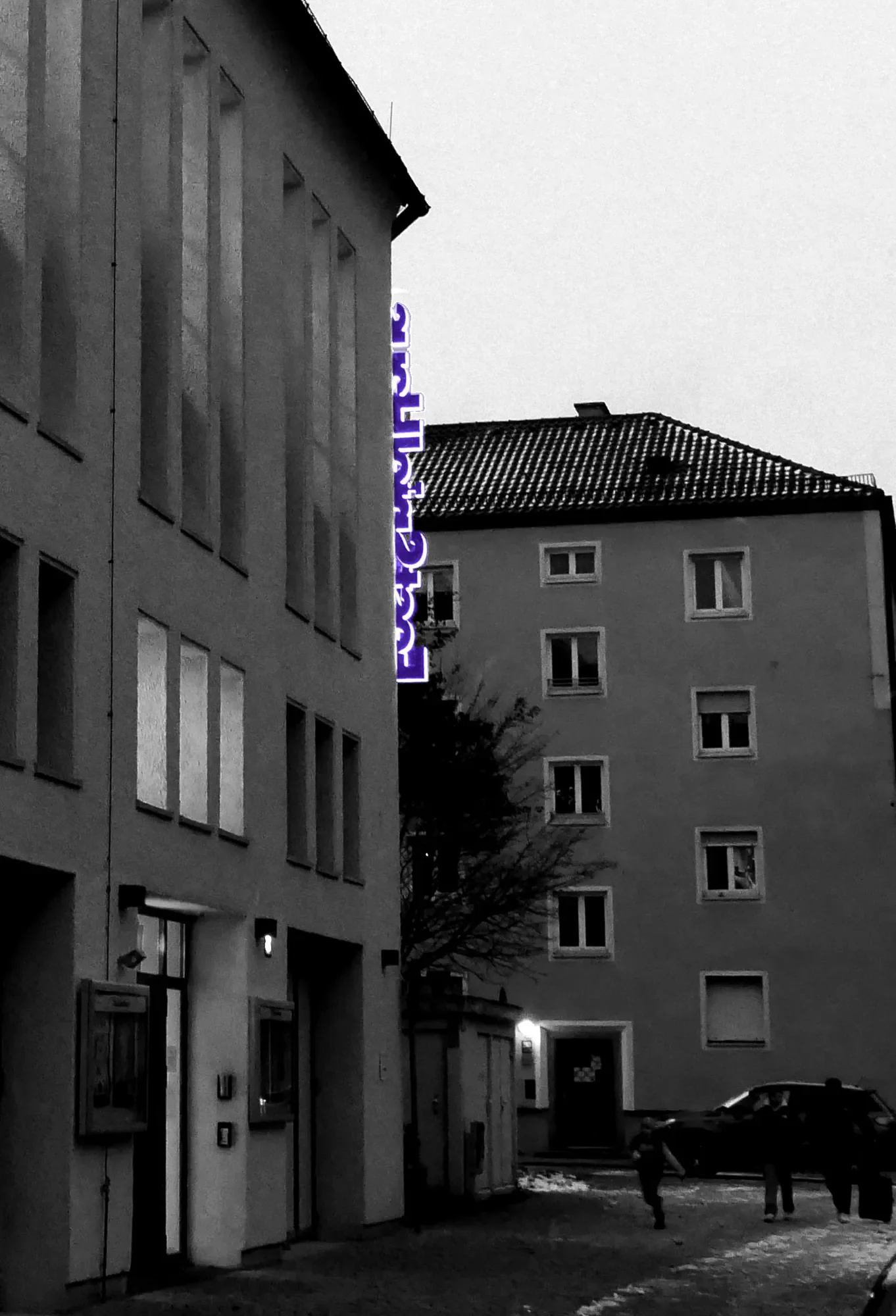 Ein schwarzes und weißes Foto einer modernen Wohnstraße bei Nacht mit einigen Menschen, die auf dem Gehweg gehen, und einem Gebäude mit einem vertikal sichtbaren Neonzeichen.