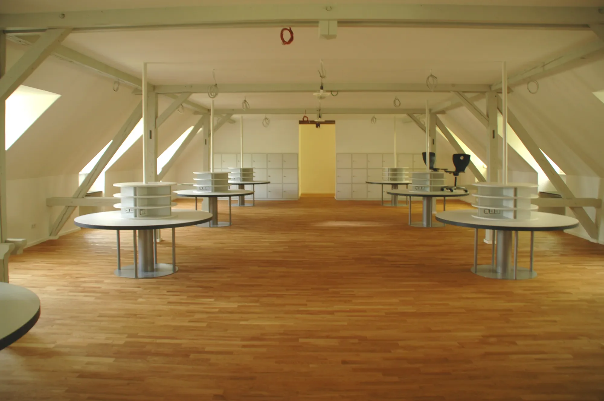 Ein leerer Raum im Dachgeschoss mit holzfarbenem Boden, weißen Wänden und mehreren runden Tischen. Es gibt mehrere Schließfächer an der Wand im Hintergrund und einige Kabel an der Decke.