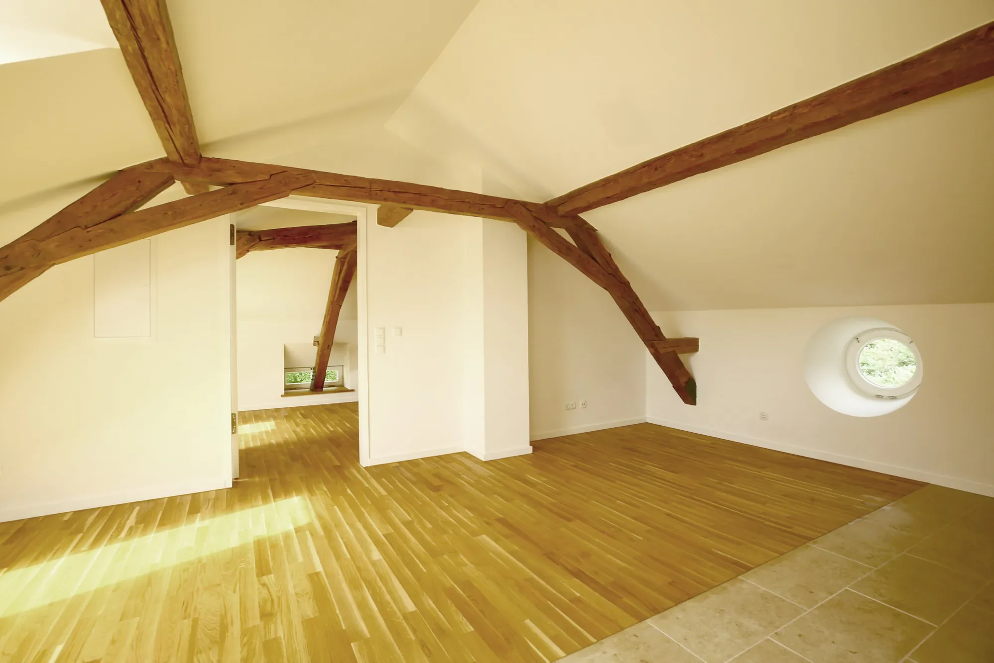 Ein heller Raum mit Holzfußboden, sichtbaren Holzbalken an der Decke, kleinen quadratischen Fenstern, einer weißen Wand und einer ovalen Dachfensters, das Tageslicht hereinlässt.