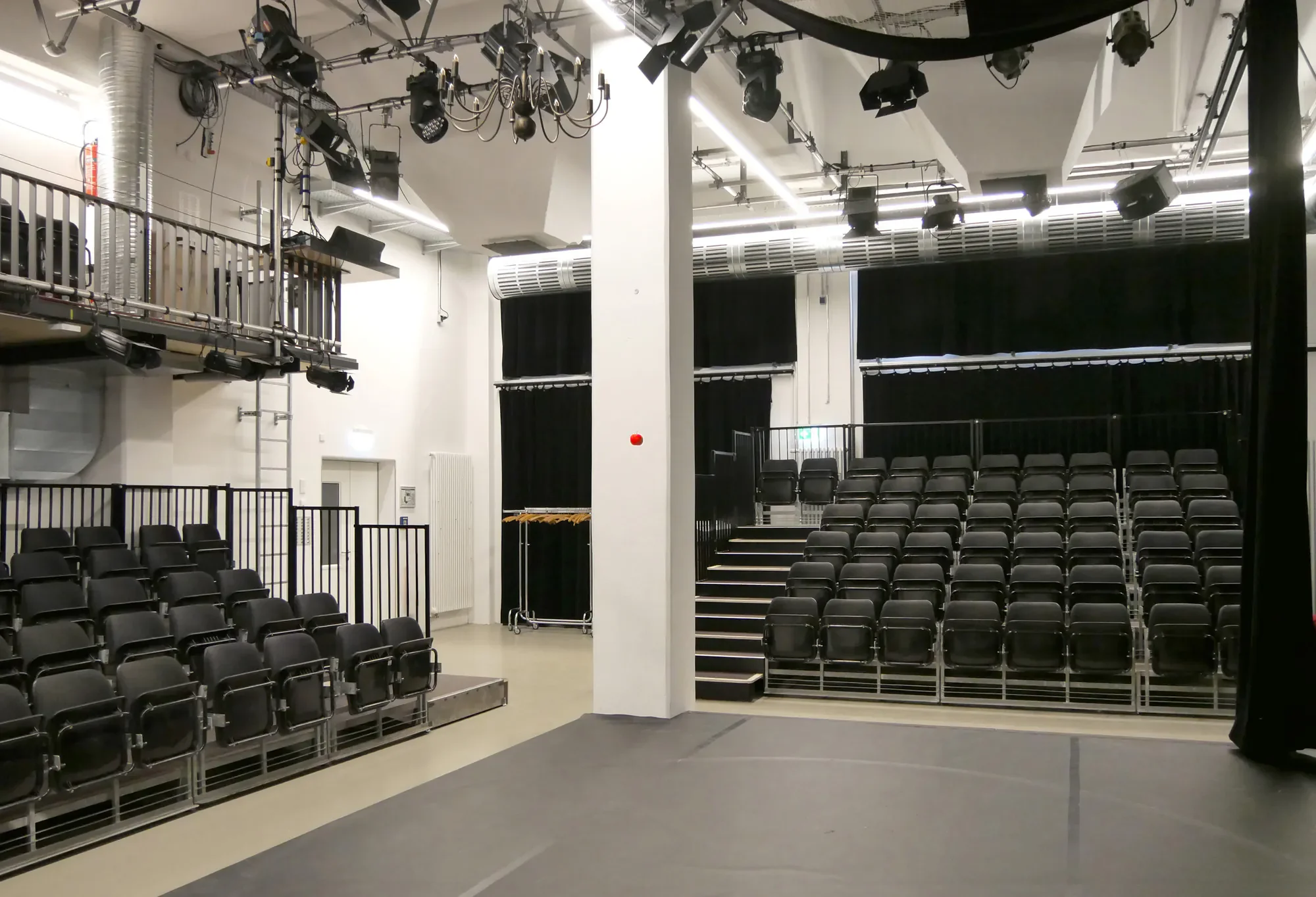Ein leeres Theater mit mehreren Reihen schwarzer Stühle, einer kleinen Bühne, schwarzem Vorhang und technischer Ausrüstung an der Decke.