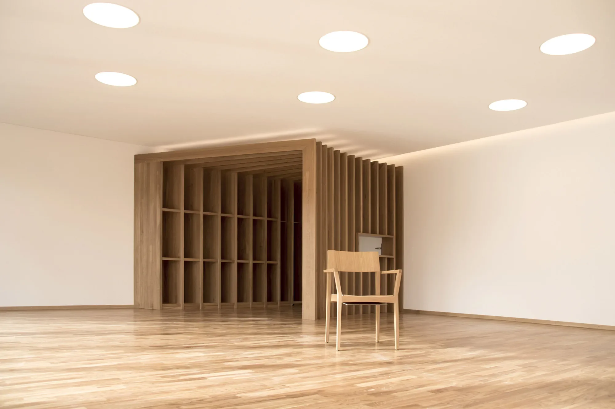 Ein moderner, minimalistischer Raum mit Holzboden, einer Holzregalwand und einem einzelnen Holzstuhl.