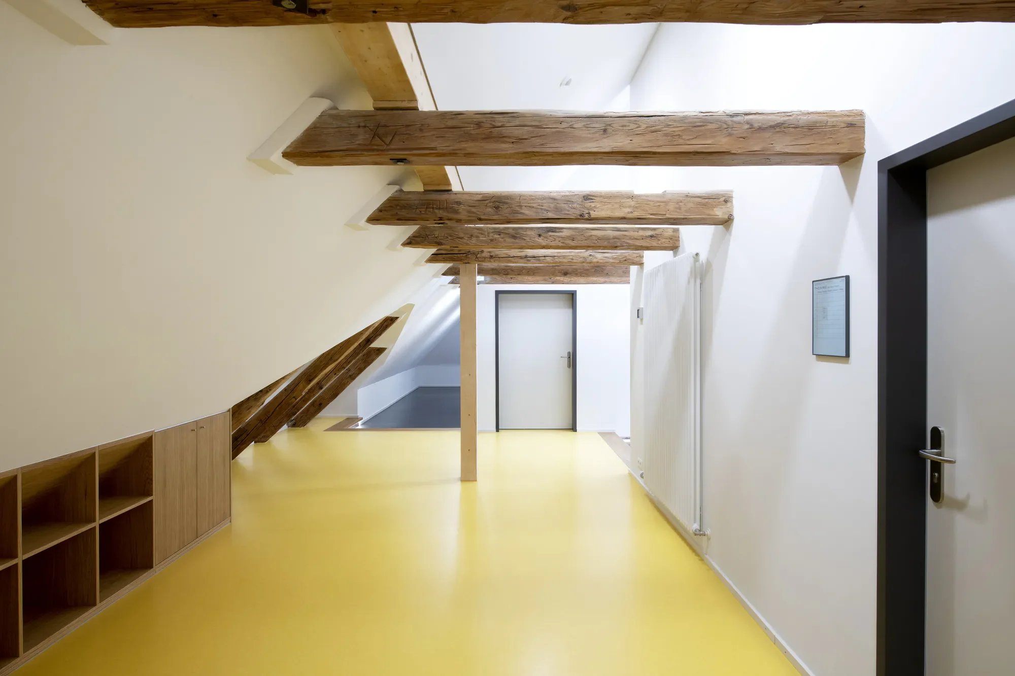 Ein heller Raum mit einer gelben Bodenfläche, weißen Wänden und sichtbaren Holzbalken an der Decke. Es gibt eine Glastür im Hintergrund, einige offene Holzregale an der linken Wand und eine Heizkörper an der rechten Wand.