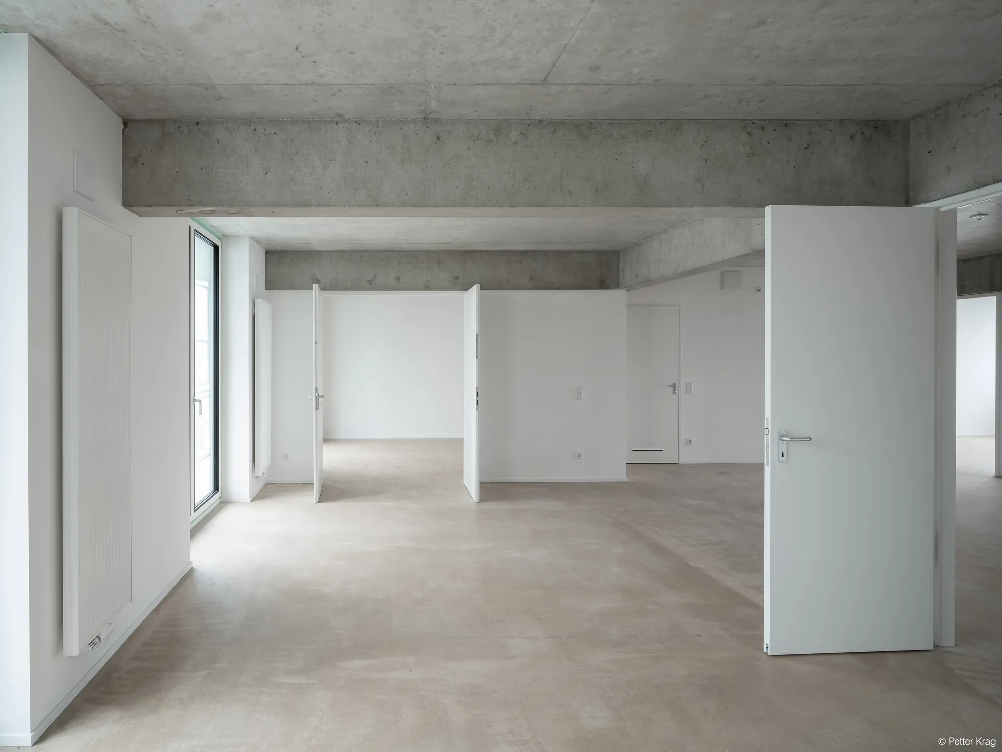Ein heller, moderner Innenraum mit weißen Wänden, großen Fenstern, offenen Türen, einem Betonboden und einer Beton-Decke.
