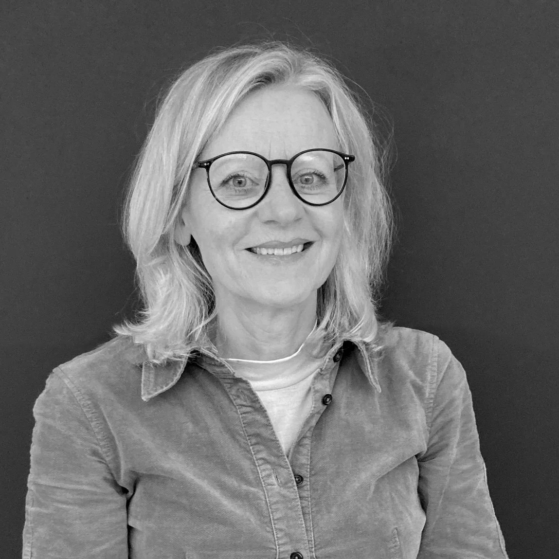 Schwarz-weiß Porträt einer Frau mit Brille, lächelnd, mit Mittellanghaar und Karohemd vor dunklem Hintergrund.