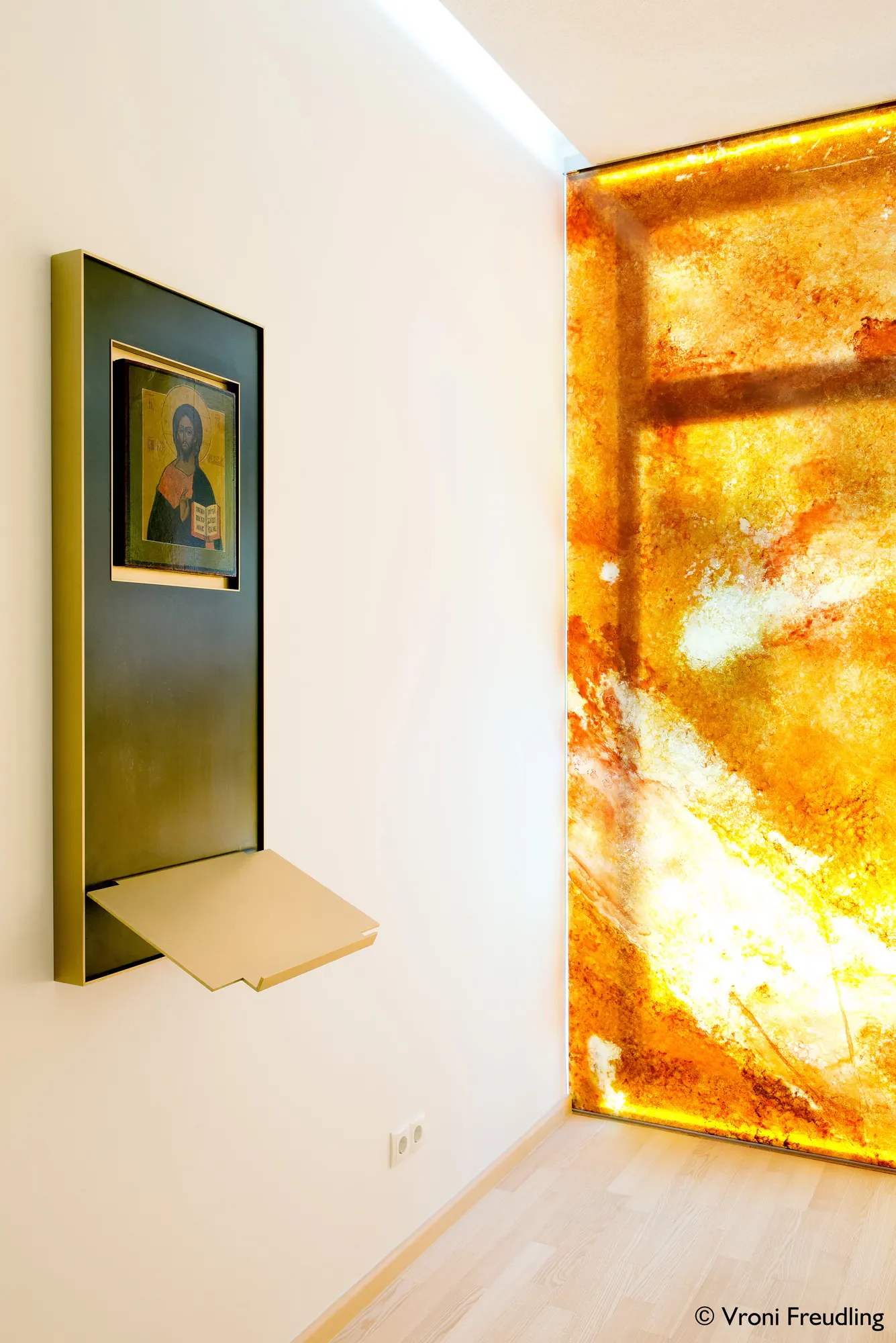 Moderner Raum mit einer weißen Wand, einem goldfarbenen Kunstwerk mit orange-gelben Farben und einem kleinen Bücherregal in Gold.