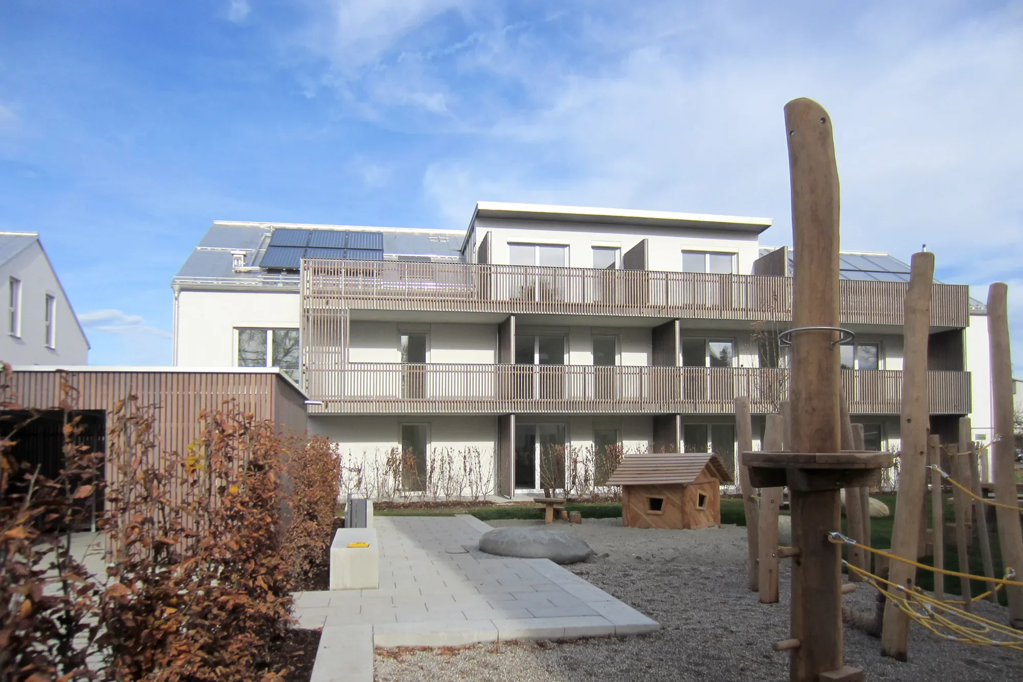 Mehrstöckiges modernes Wohngebäude mit großen Fenstern und Holzgeländer auf dem Balkon, Solarzellen auf dem Dach, im Vordergrund Spielplatz mit Baumhaus und Holzpferd, blauer Himmel mit wenigen Wolken.