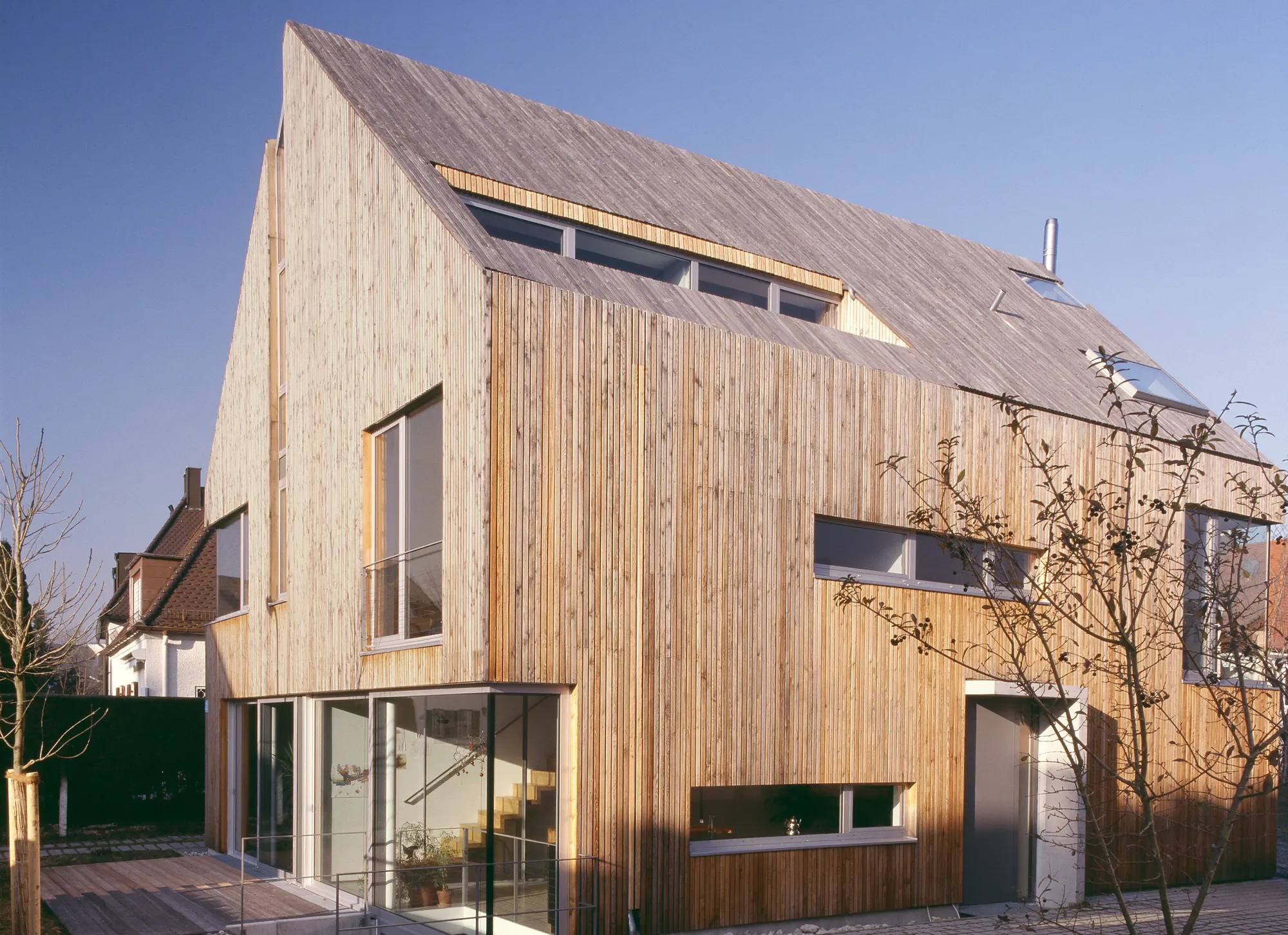 Modernes mehrstöckiges Haus mit Holzfassade und großen Fensterflächen; Dächer mit Dachfenstern und schrägen Flächen; Bäume im Vordergrund bei sonnigem Himmel.