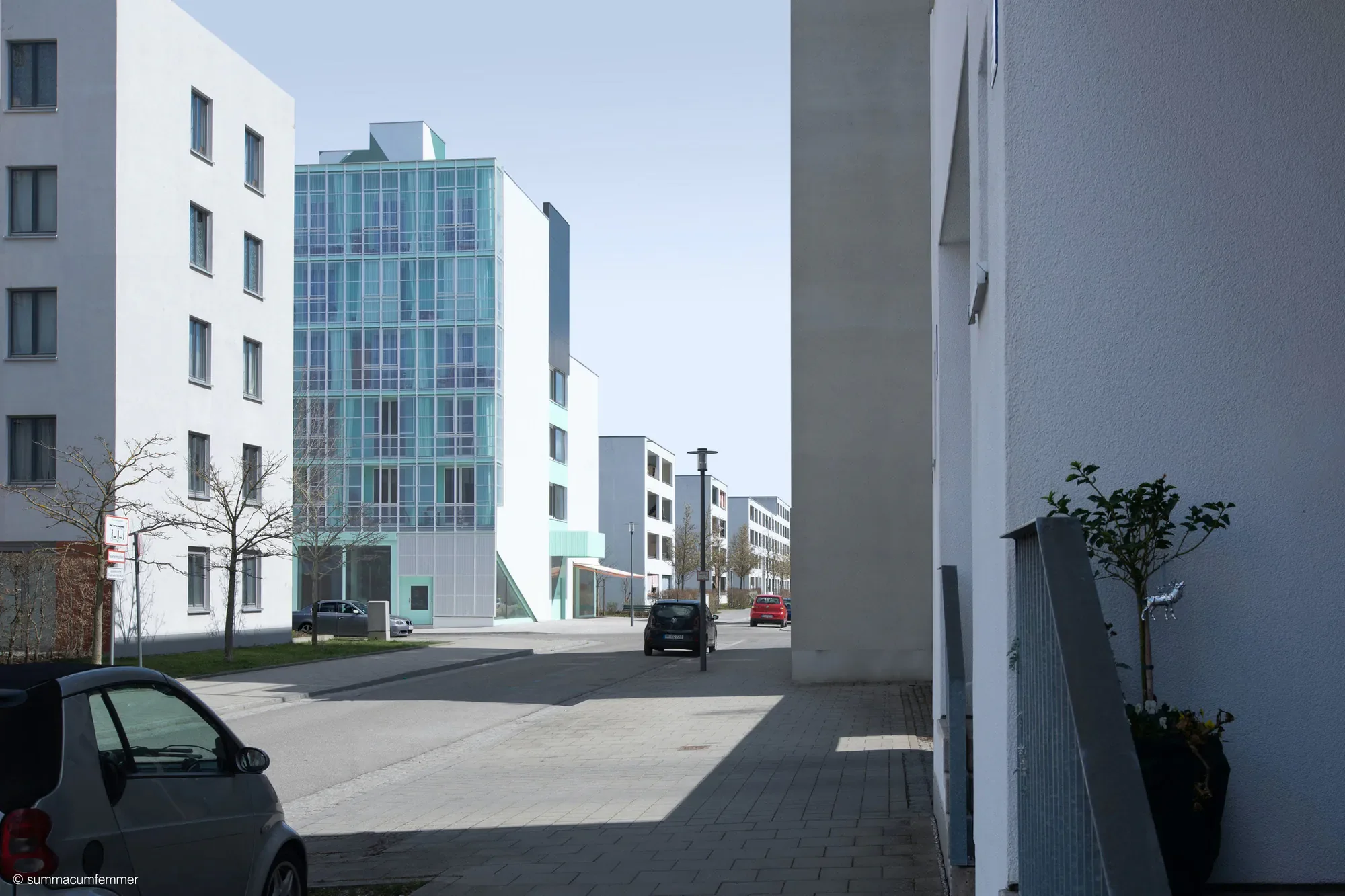 Ein Blick auf eine moderne Stadtstraße mit weißen Gebäuden, einem gläsernen Hochhaus im Hintergrund, einigen Bäumen, parkenden Autos und Straßenlaternen bei sonnigem Wetter.