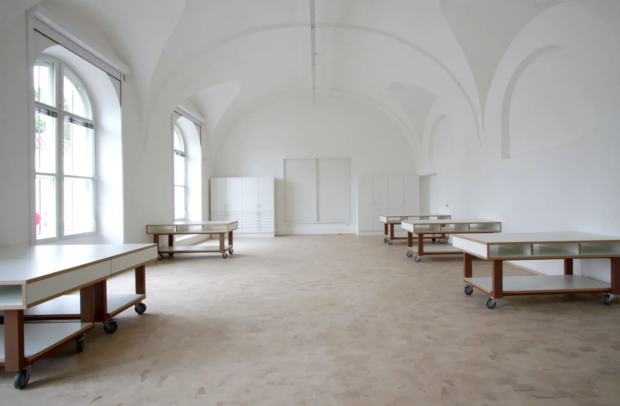 Ein heller, leerer Raum mit großen, arched Fenstern und weißer Wände, eingerichtet mit rollbaren Tischen.