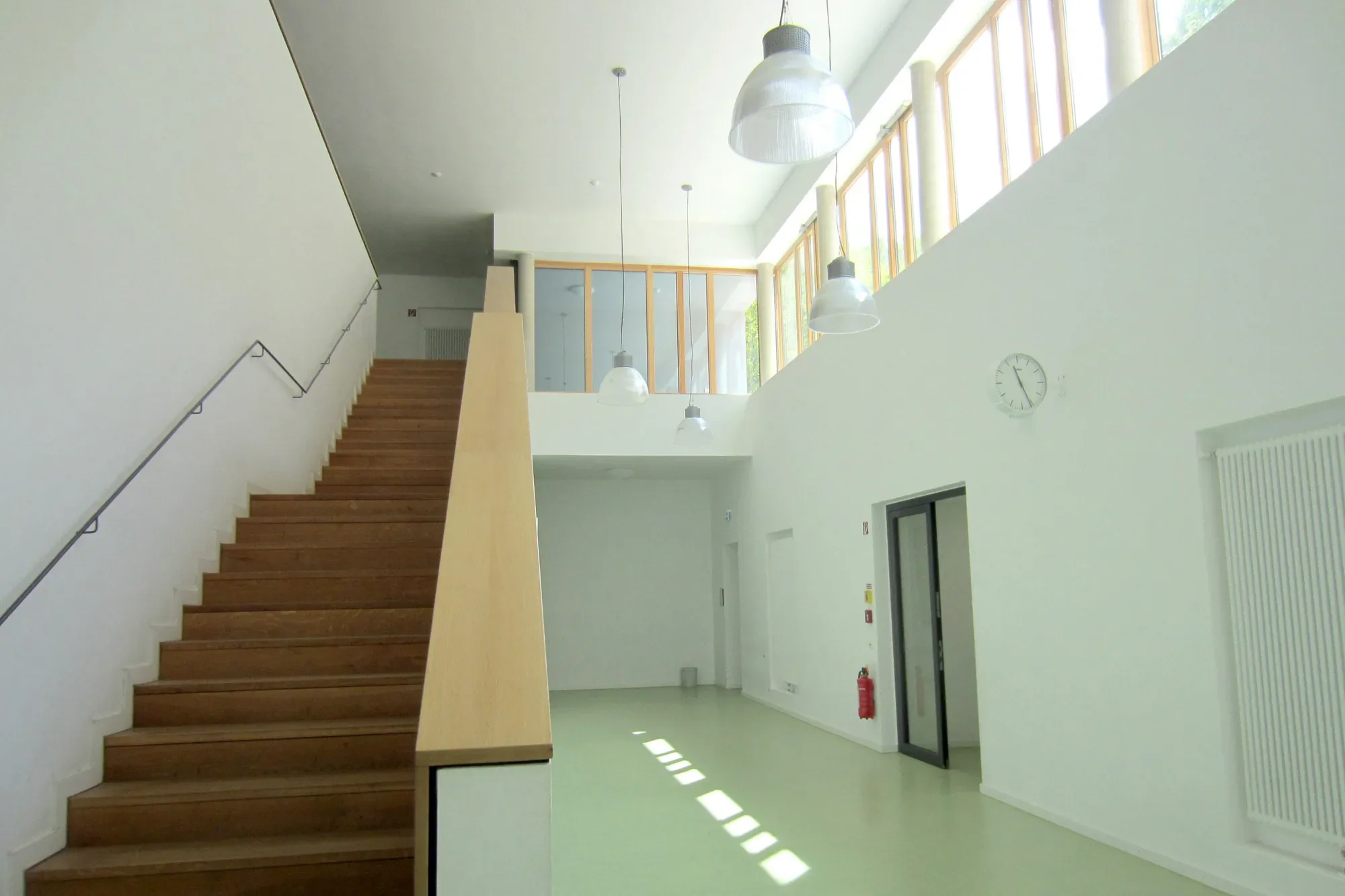 Innenansicht eines modernen Gebäudes mit Treppe aus Holz, großen Fenstern, weißen Wänden und mehreren Hängeleuchten. Es gibt eine Uhr an der Wand und eine Feuerlöscherstation auf dem Flur.