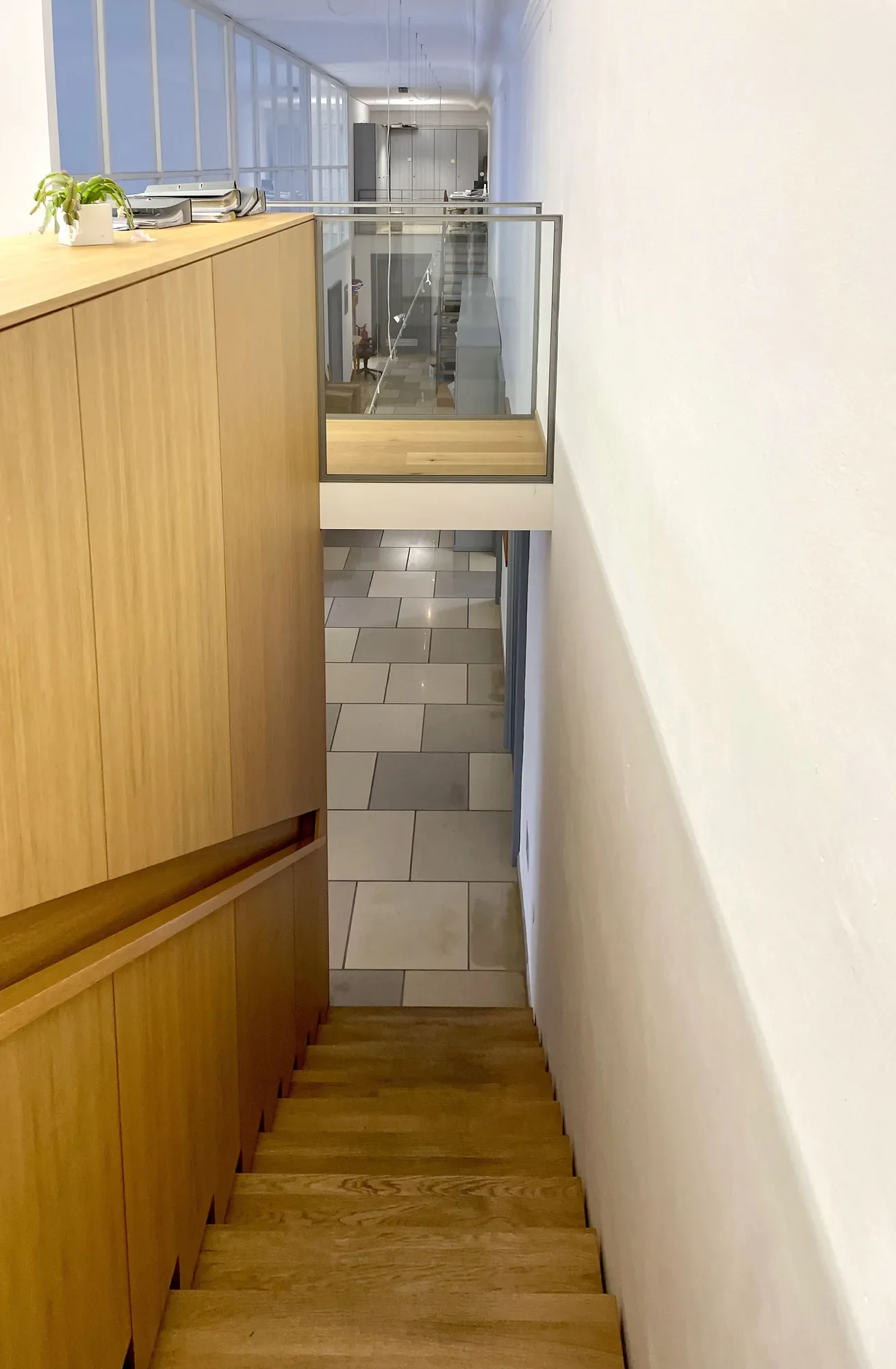 Innenansicht eines Treppenhauses mit Holzsichttreppe und Glasgeländer, daneben eine weiße Wand, im Hintergrund Büros mit großen Fenstern.