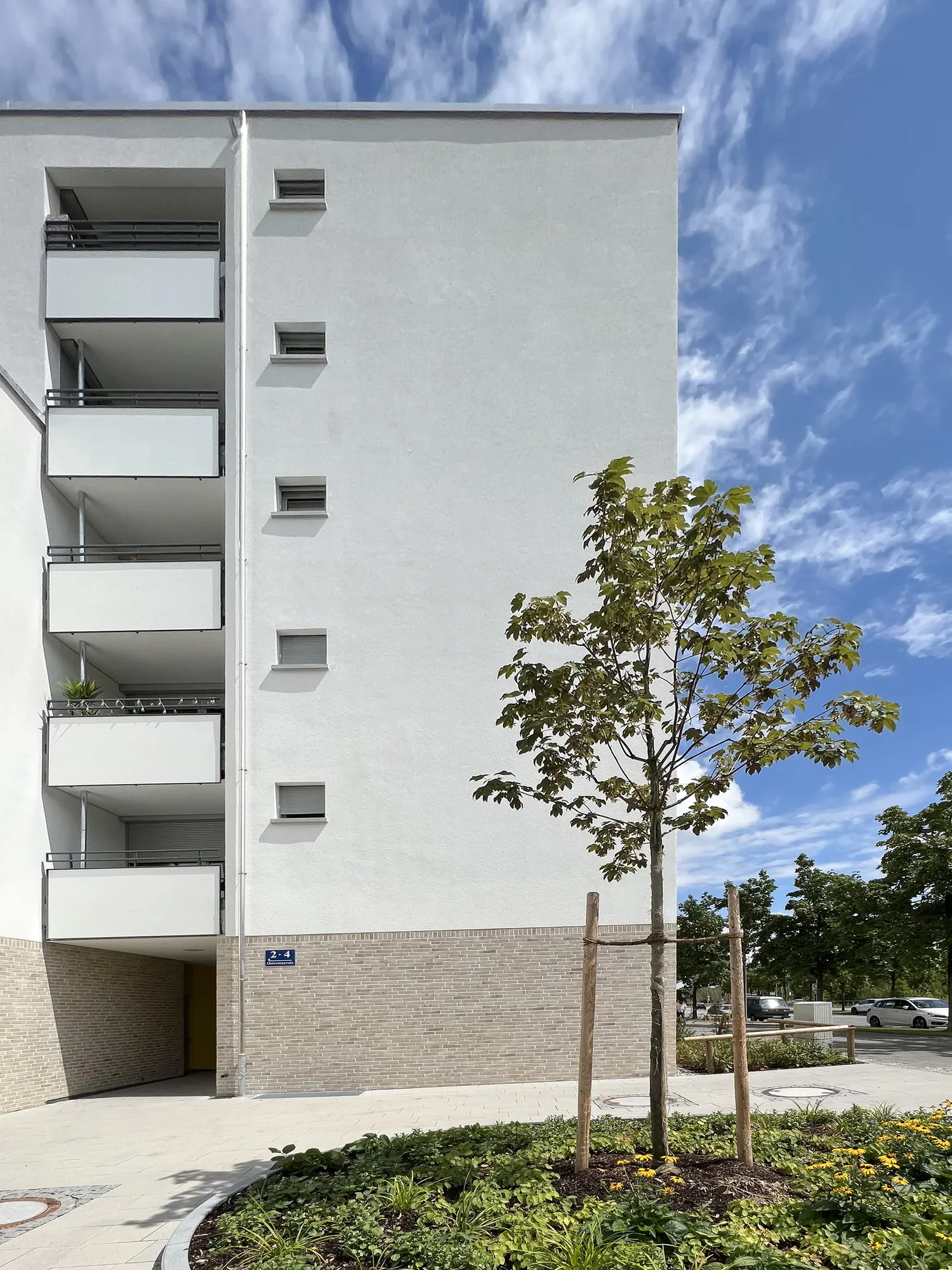 Mehrstöckiges modernes Wohngebäude mit Balkonen und kleinen Dachfenstern an der Fassade, Bäume im Vordergrund, blauer Himmel mit Wolken, Straßenverkehr im Hintergrund.