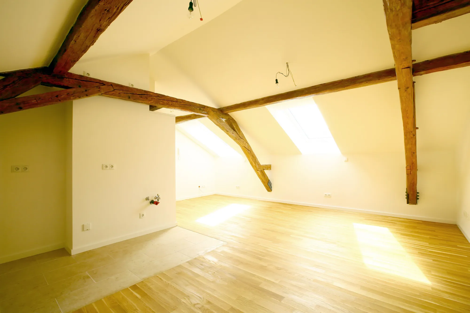 Ein heller Raum mit Holzbalken an der Decke und großen Dachfenstern, leeres Laminatboden, keine Möbel, Kabel hängen von der Decke.