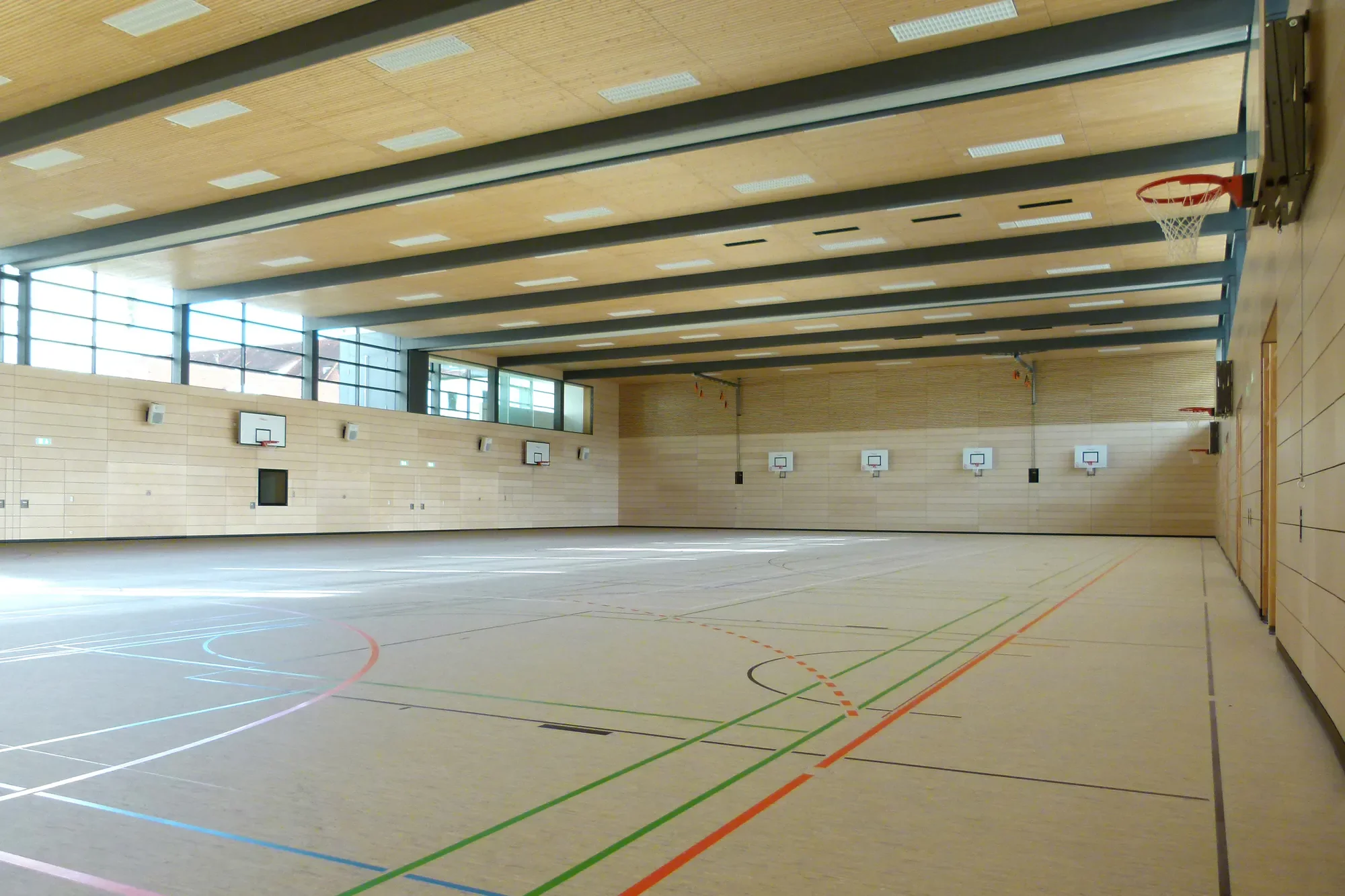 Leerer Turnhalle mit Basketballkörben an den Wänden, großen Fenstern, hölzernem Deckenboden und verschiedenen Linien für Sportarten auf dem Boden.