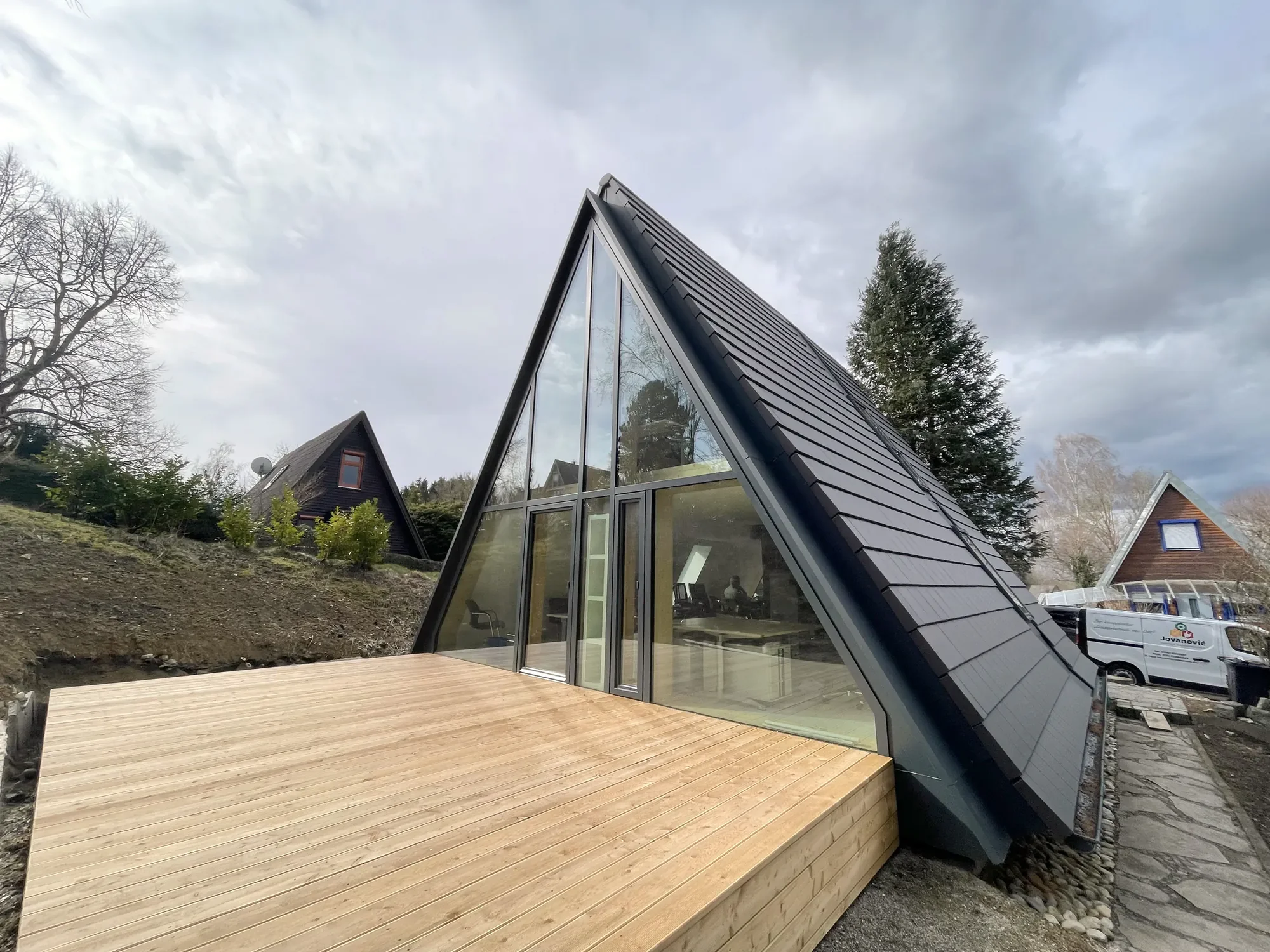 Modernes Haus mit großen Glasflächen und schrägem schwarzen Dach, umgeben von Bäumen und weiteren Häusern.