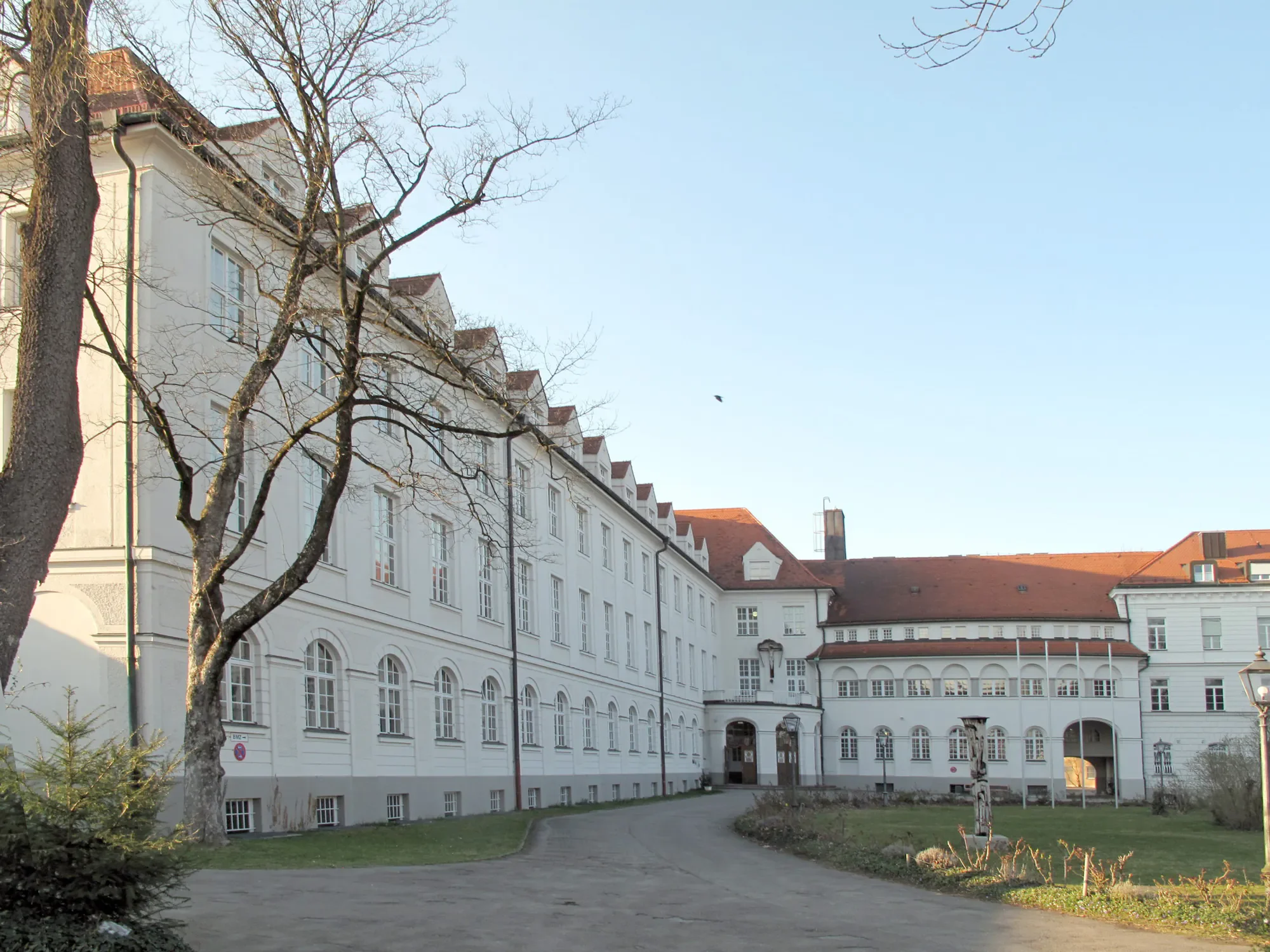 Ein weißes Gebäude mit roten Dächern und mehreren Fenstern im Hintergrund, Bäume im Vordergrund, blauer Himmel.