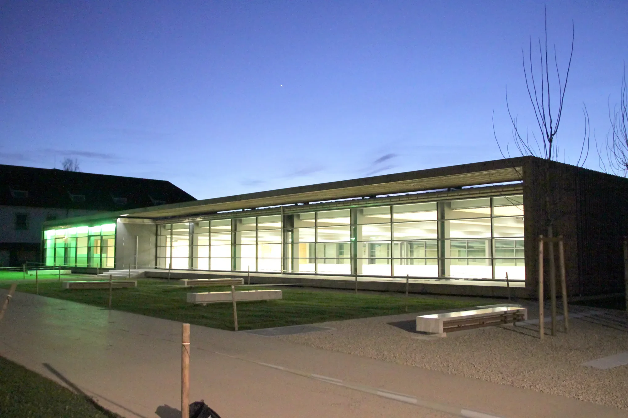 Ein modernes Gebäude mit großen Glasfenstern, das nachts beleuchtet ist, im Hintergrund ein blauer Himmel und eine kleine helle Stern. Im Vordergrund ein Gehweg, einige Bänke und eine unbepflanzte Fläche.