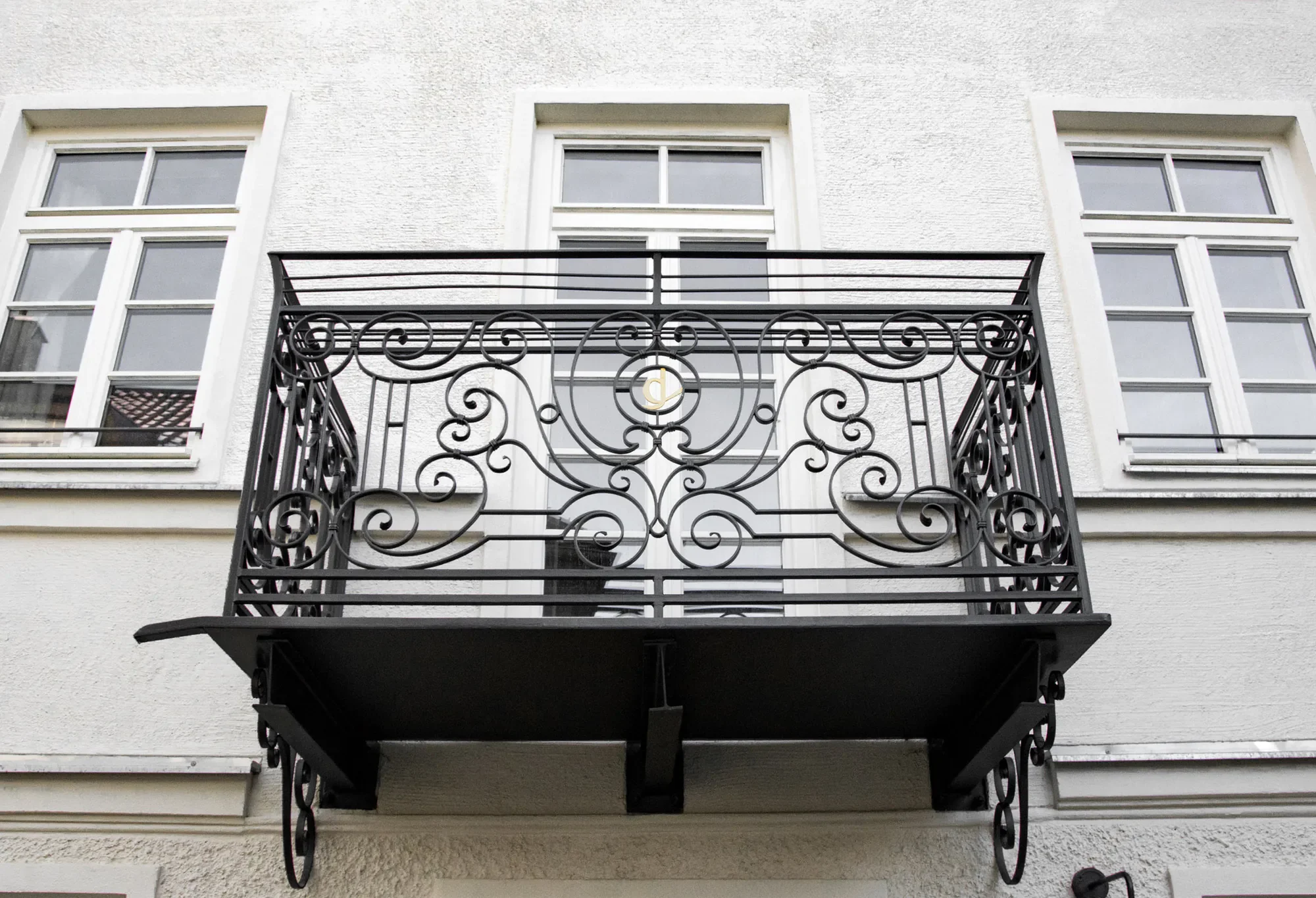 Balkon mit filigranem schwarzen Schmiedeeisen-Geländer an einer weißen Fassadenwand, darüber drei Fenster